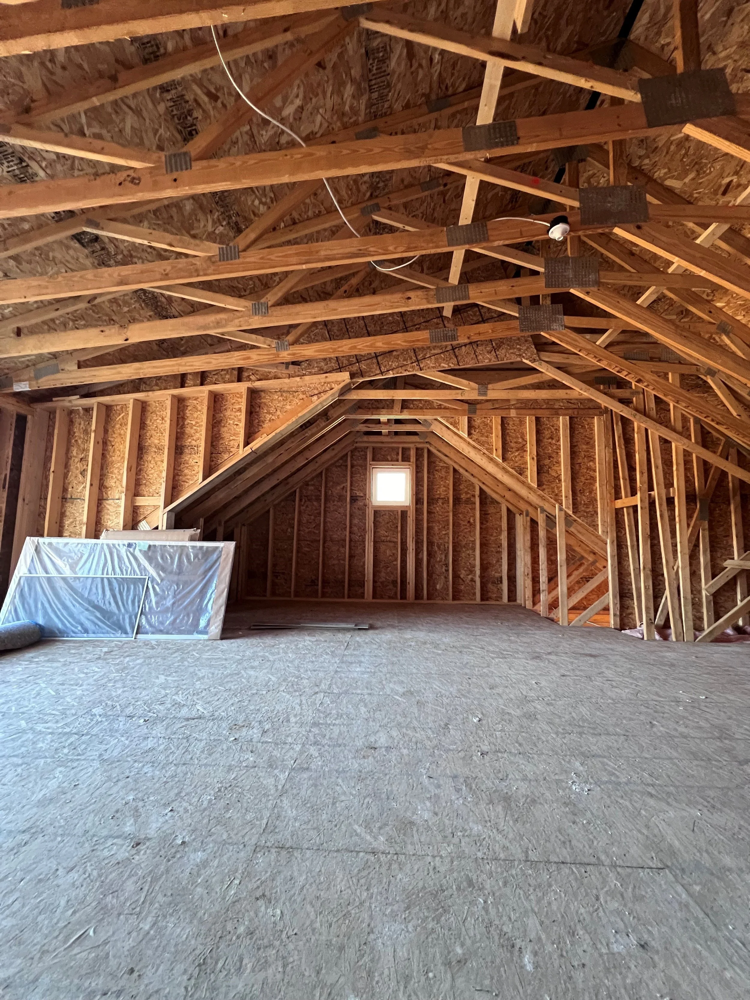 Interior view of an unfinished attic with exposed wooden roof trusses and plywood sheathing, with a small window at the end and construction materials on the floor.
