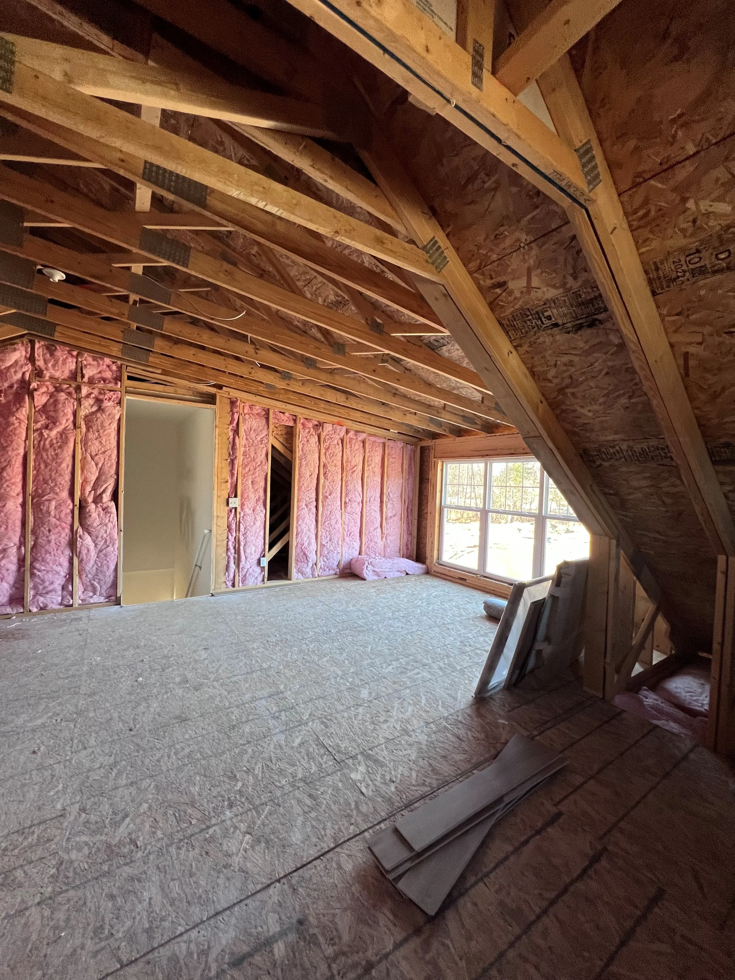 Interior of a room under construction with exposed insulation and wooden framing. There are construction materials and tools visible, with a window letting in natural light.