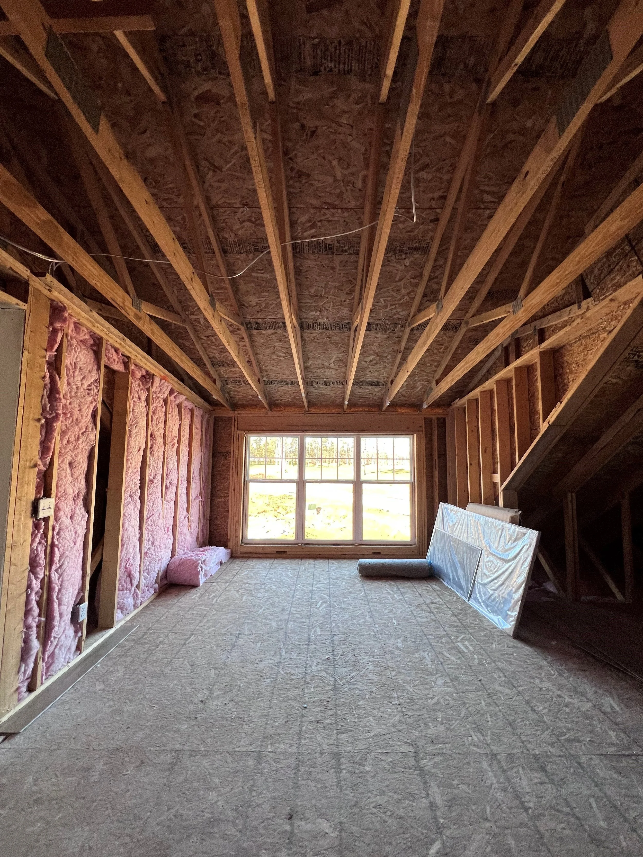 Interior of a room under construction, with exposed wooden studs, pink insulation on the walls, a window, and construction materials on the floor.