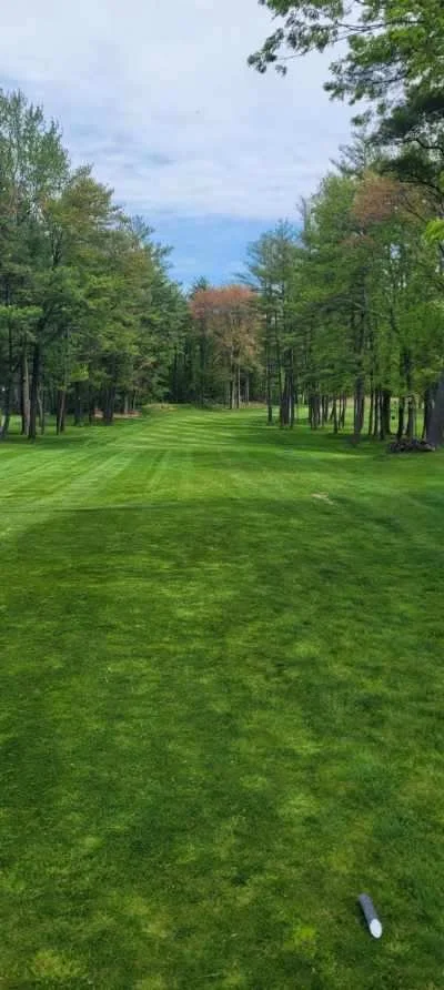 A golf course fairway with green grass, surrounded by trees on both sides, and a partly cloudy sky above.