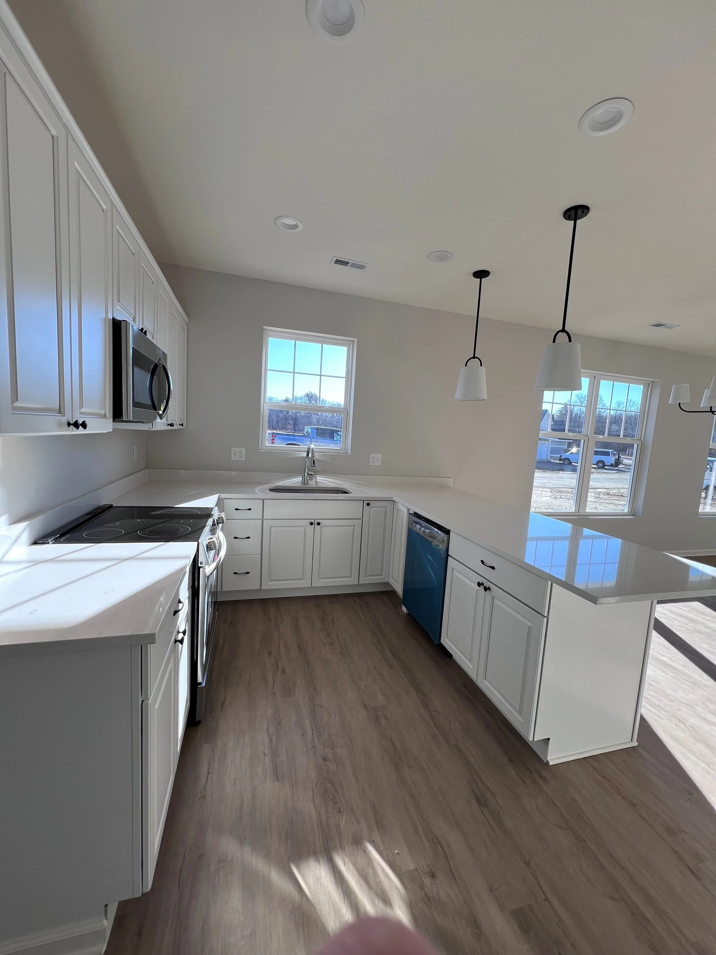 Bright kitchen with white cabinets, stainless steel appliances, two windows, pendant lights, and a wide island with a white countertop.