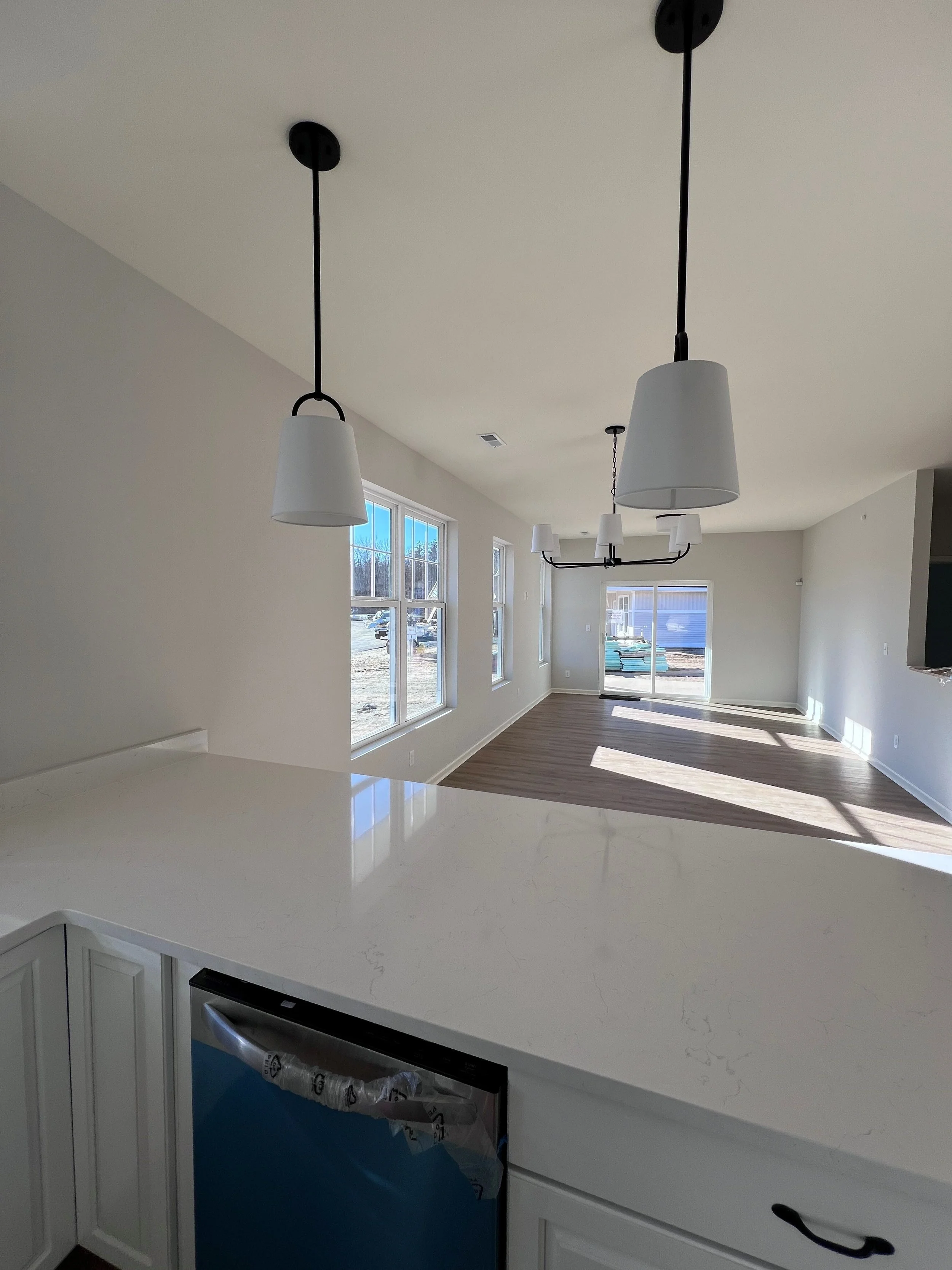 Modern kitchen with a white countertop, white cabinets, sunlight streaming in through large windows, and ceiling lights hanging above.
