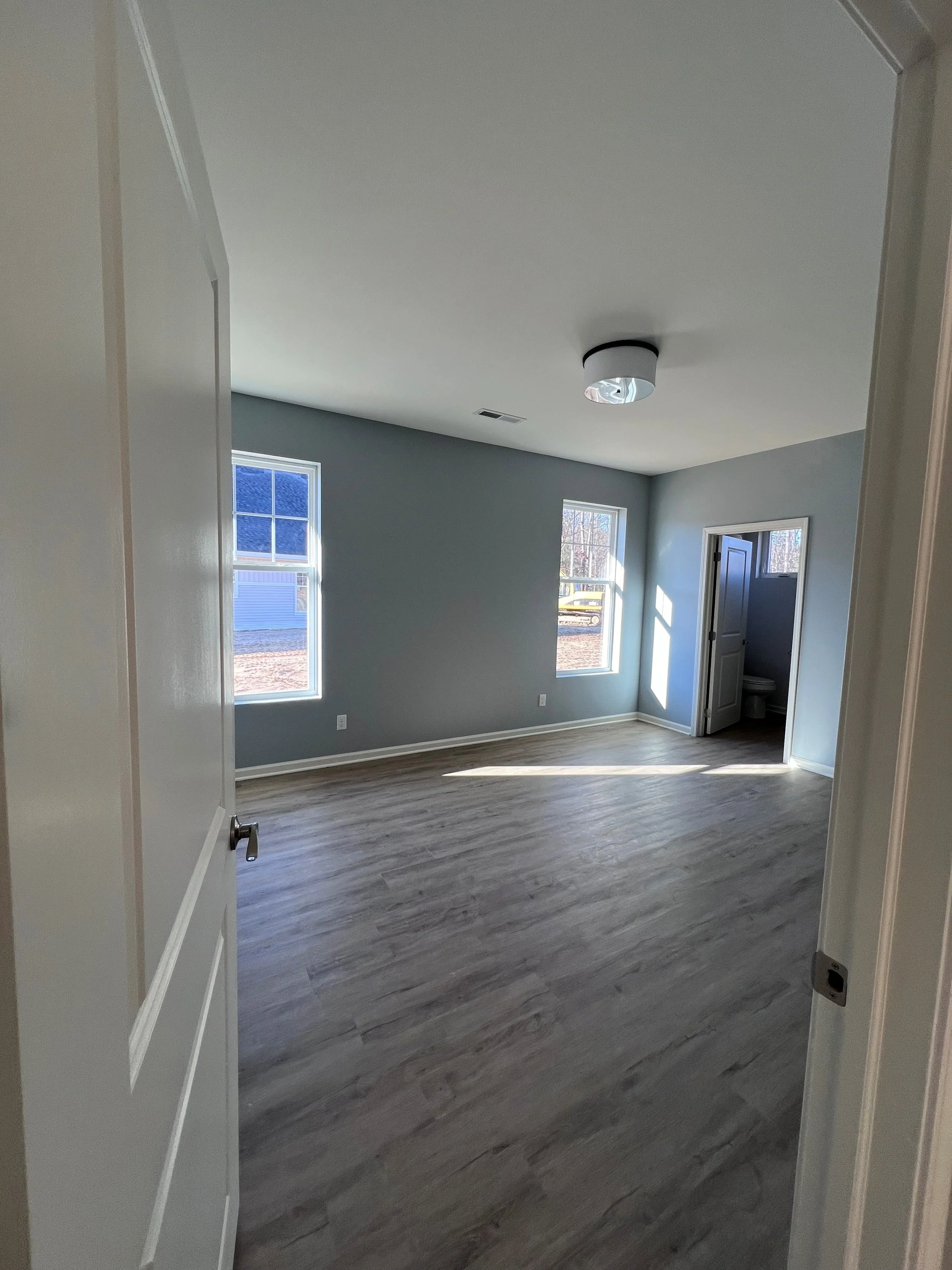 Empty room with large windows, gray walls, and wood-style flooring. The room has a ceiling light fixture and a door leading to a bathroom.