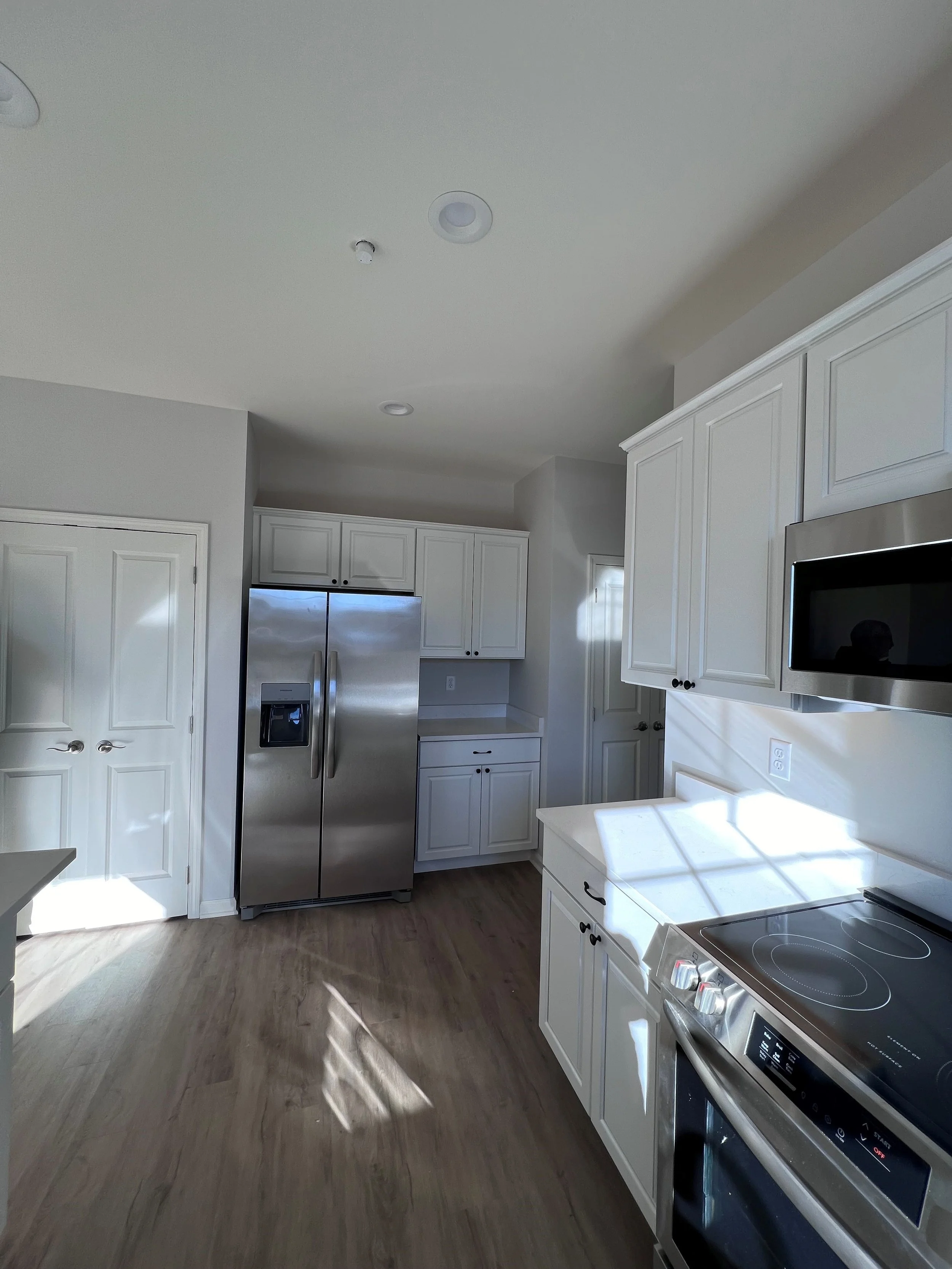 A modern kitchen with white cabinets, a stainless steel refrigerator, a microwave, and a stove, with sunlight casting shadows on the wooden floor.