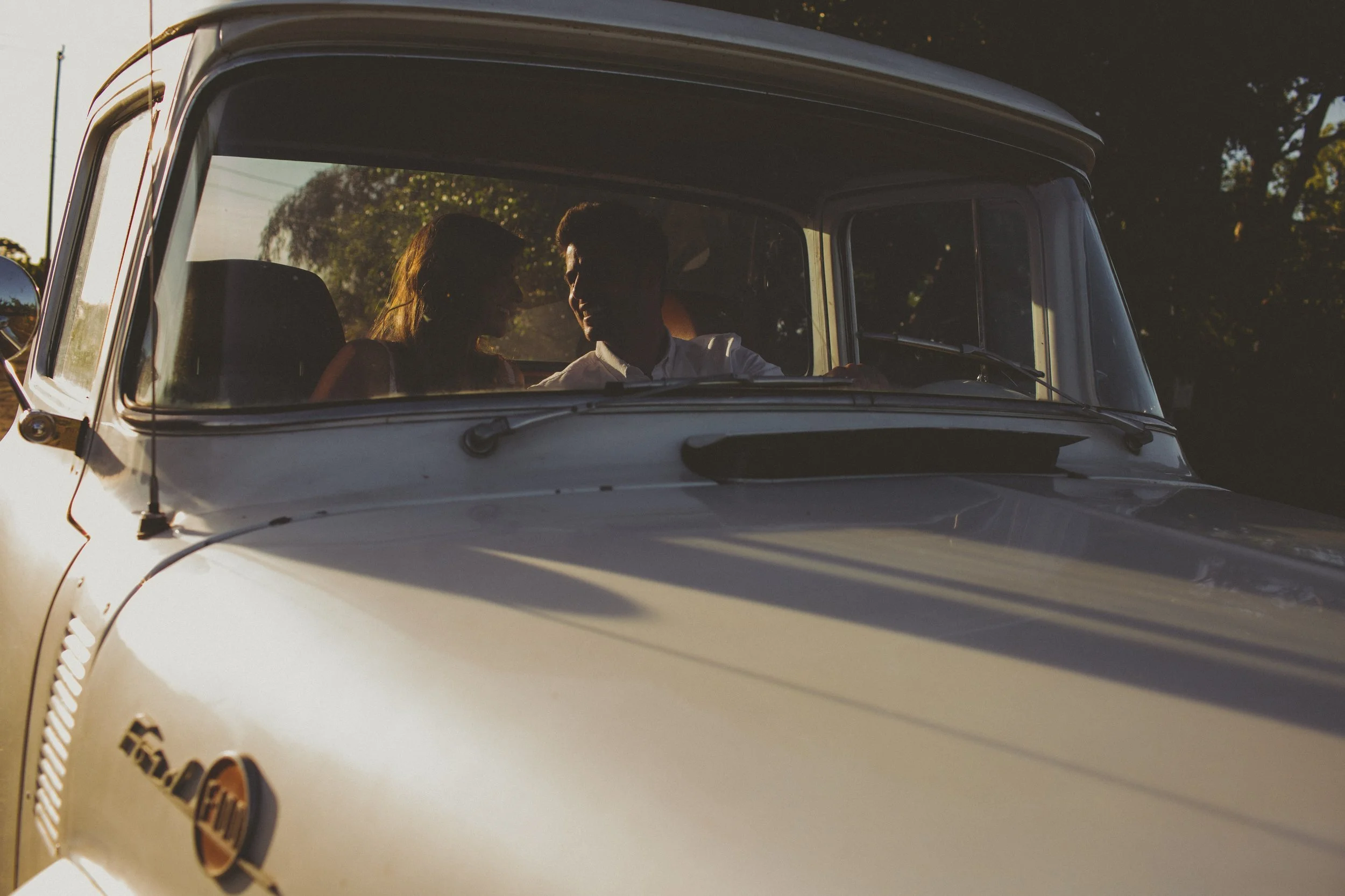 A couple sitting inside a vintage white truck, silhouetted against the sunset outside, engaging in conversation.