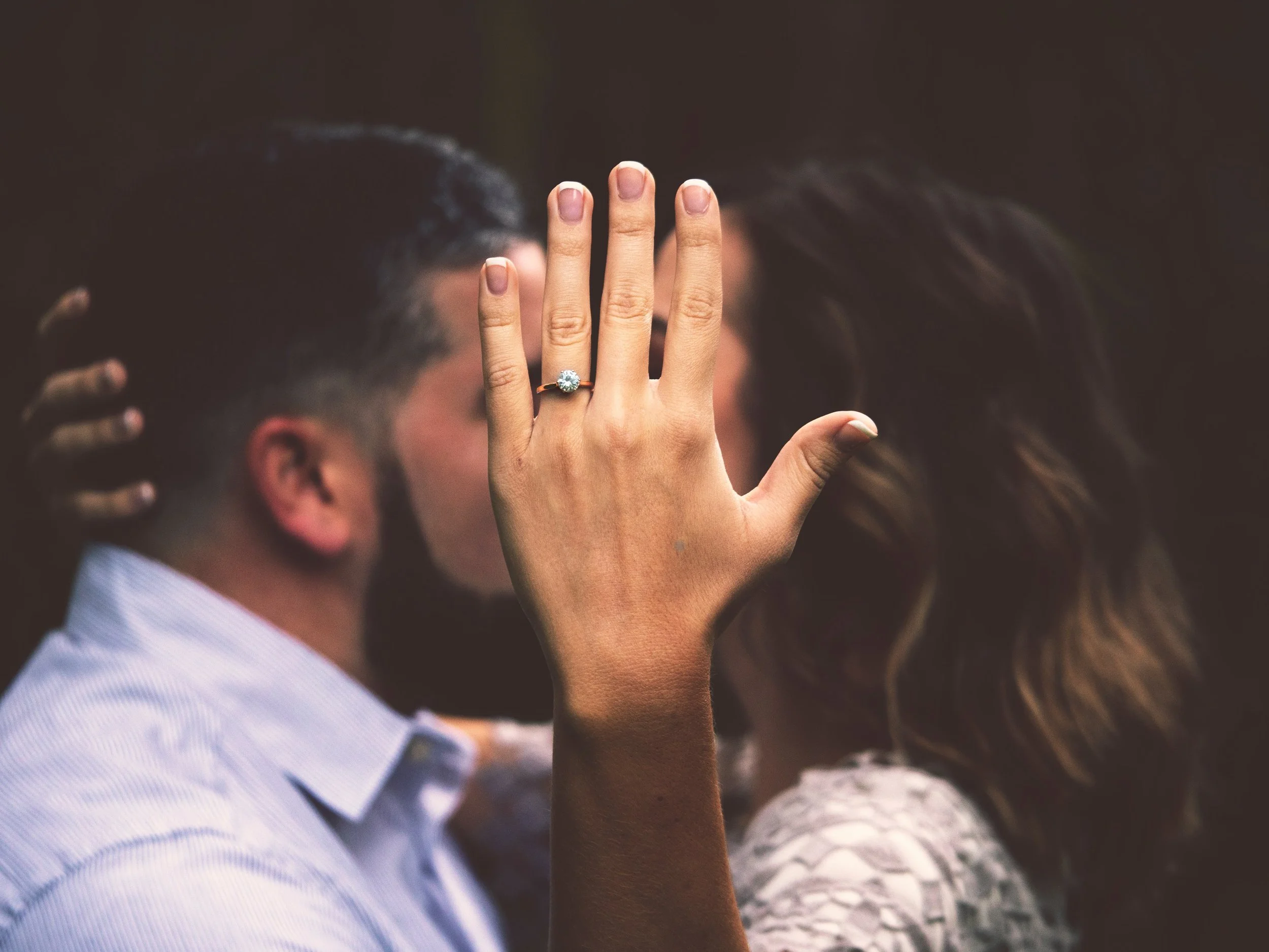 Close-up of a woman's hand with an engagement ring, covering the face of a man, as they share a kiss in the background.