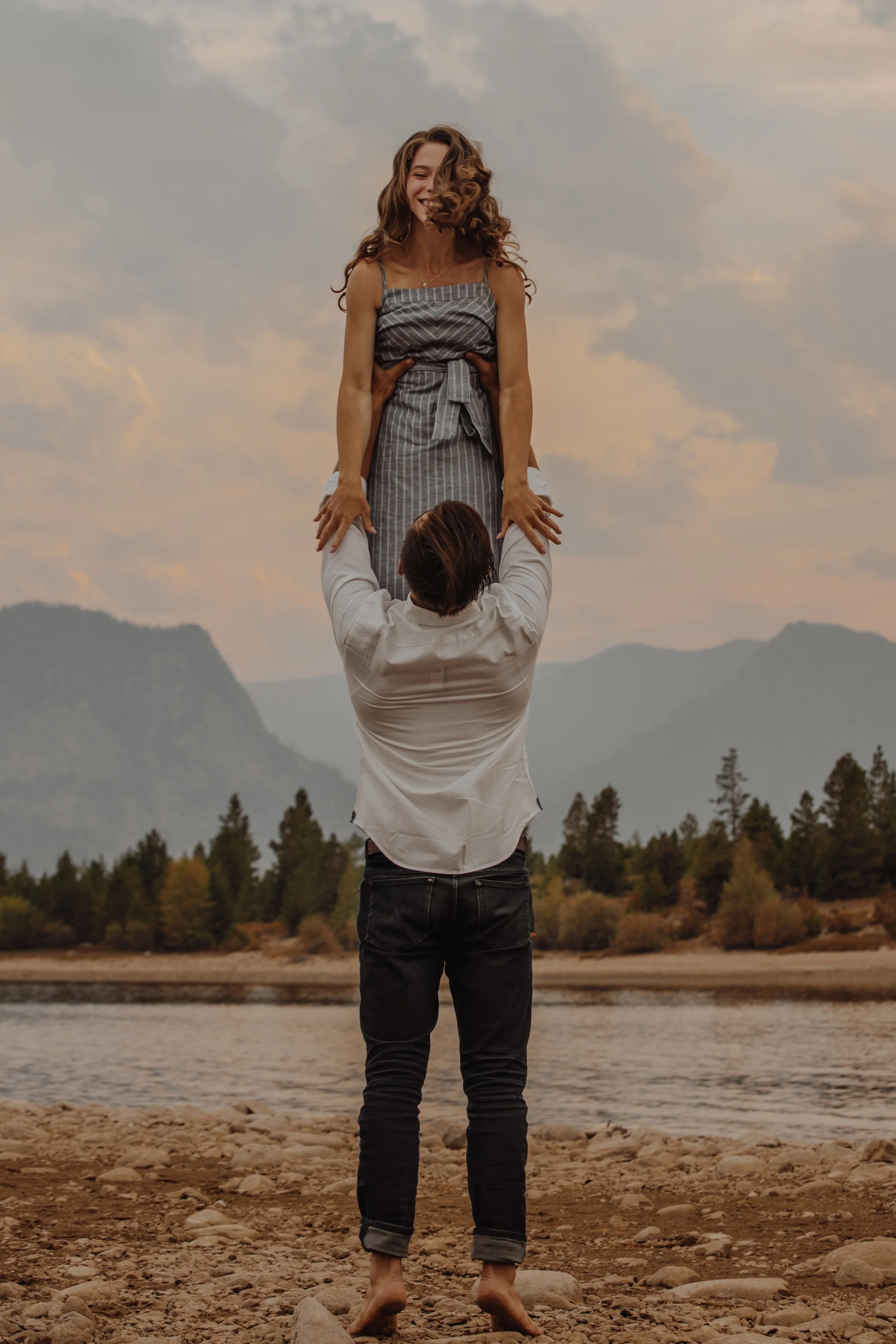 A man is holding a woman up above his head with both hands while standing barefoot on a rocky shore, with mountains, trees, and a cloudy sky in the background.