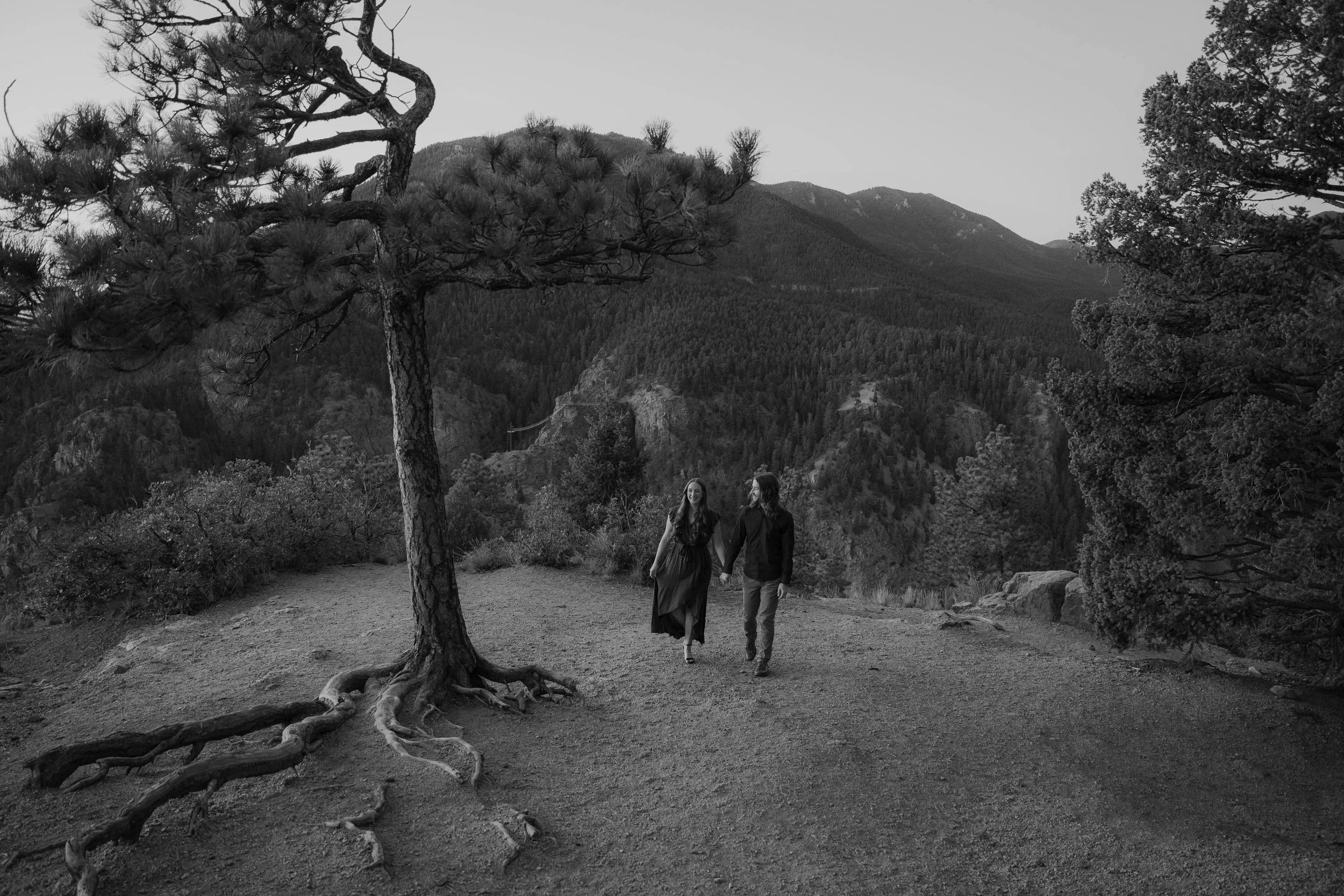 A black and white photo of a couple walking hand in hand on a dirt trail in a mountainous forest.
