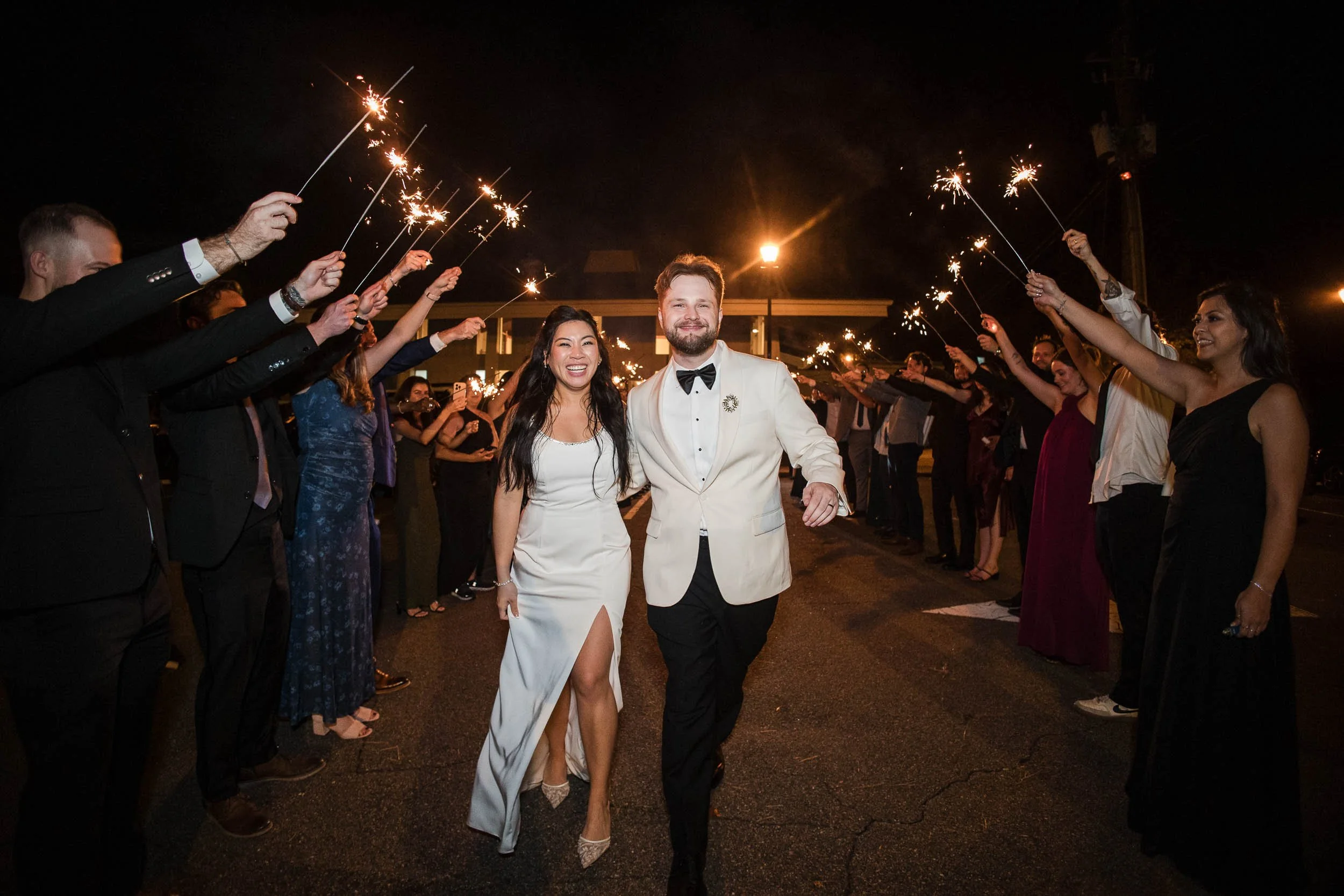 A couple in formal wedding attire walking through a crowd holding sparklers at night.