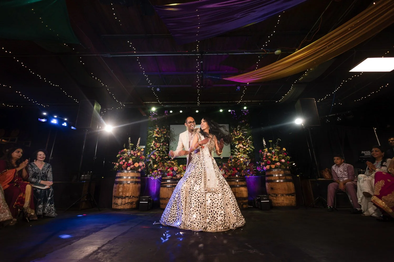 An Indian couple dancing at their Sangeet reception, surrounded by guests and floral decorations, with colorful draped fabric and string lights hanging from the ceiling at Wild Heaven Beer Room Venue.