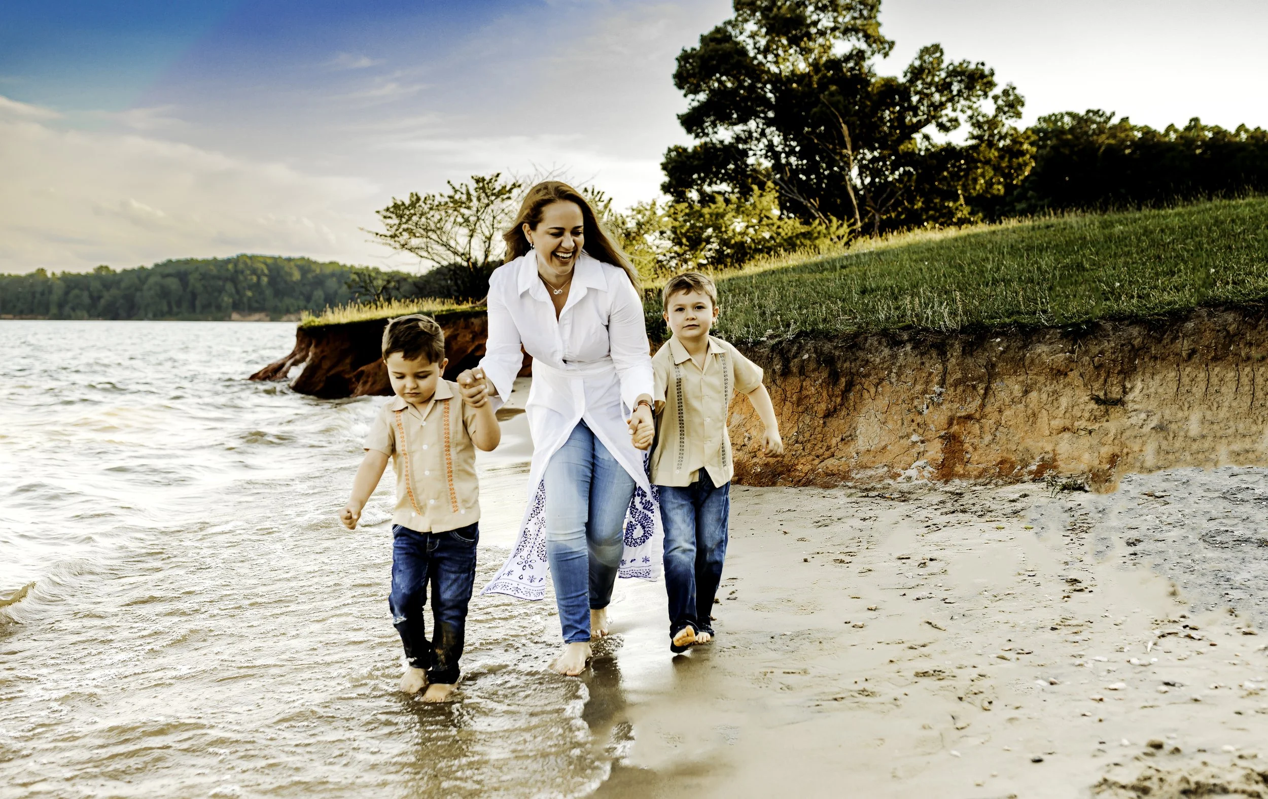 Woman and two young boys walking hand-in-hand along the shoreline of a lake or river during sunset.