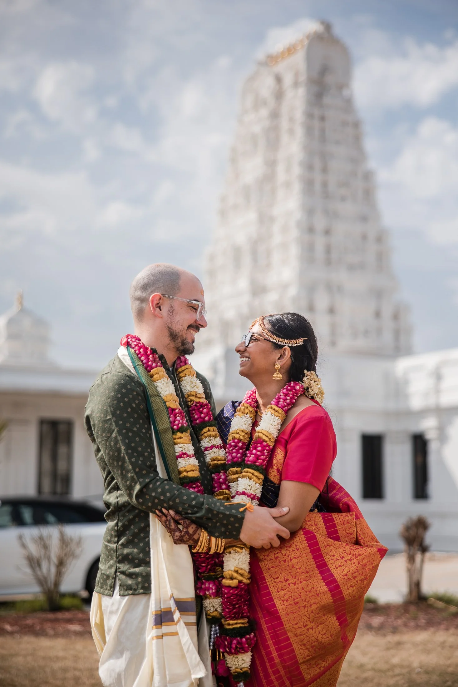 A couple dressed in traditional Indian attire, wearing floral garlands, standing outdoors in front of a white temple, smiling at each other.