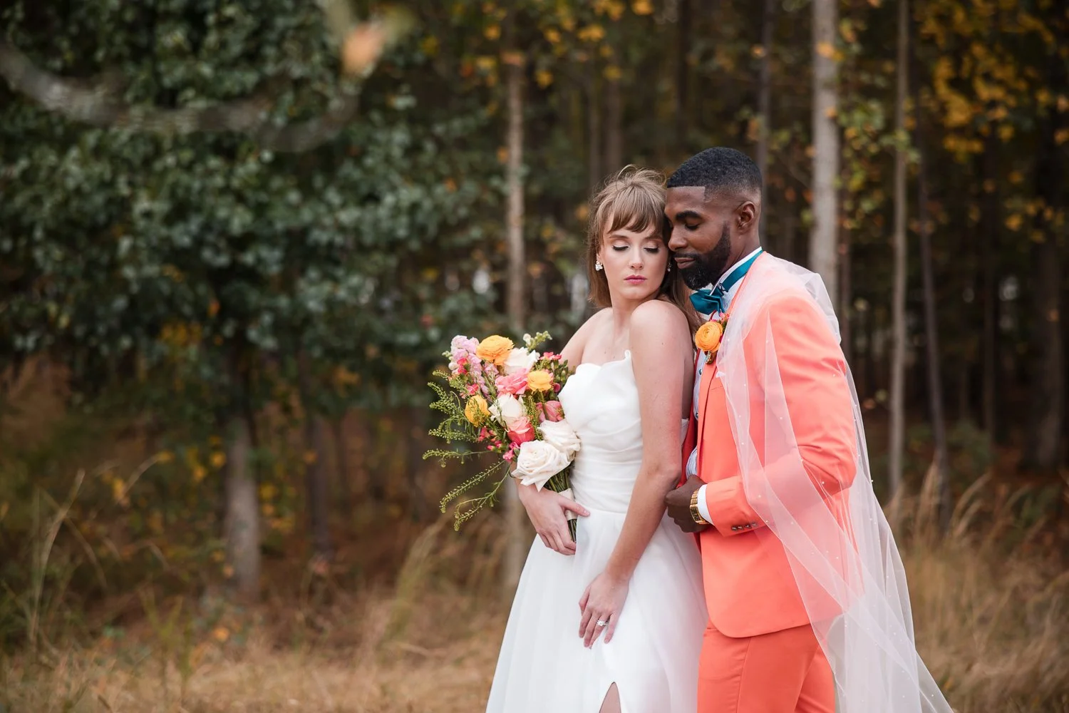 A bride in a white wedding dress holding a colorful bouquet of flowers next to a groom in an orange suit with a veil draped over his shoulder, standing outdoors with trees in the background.