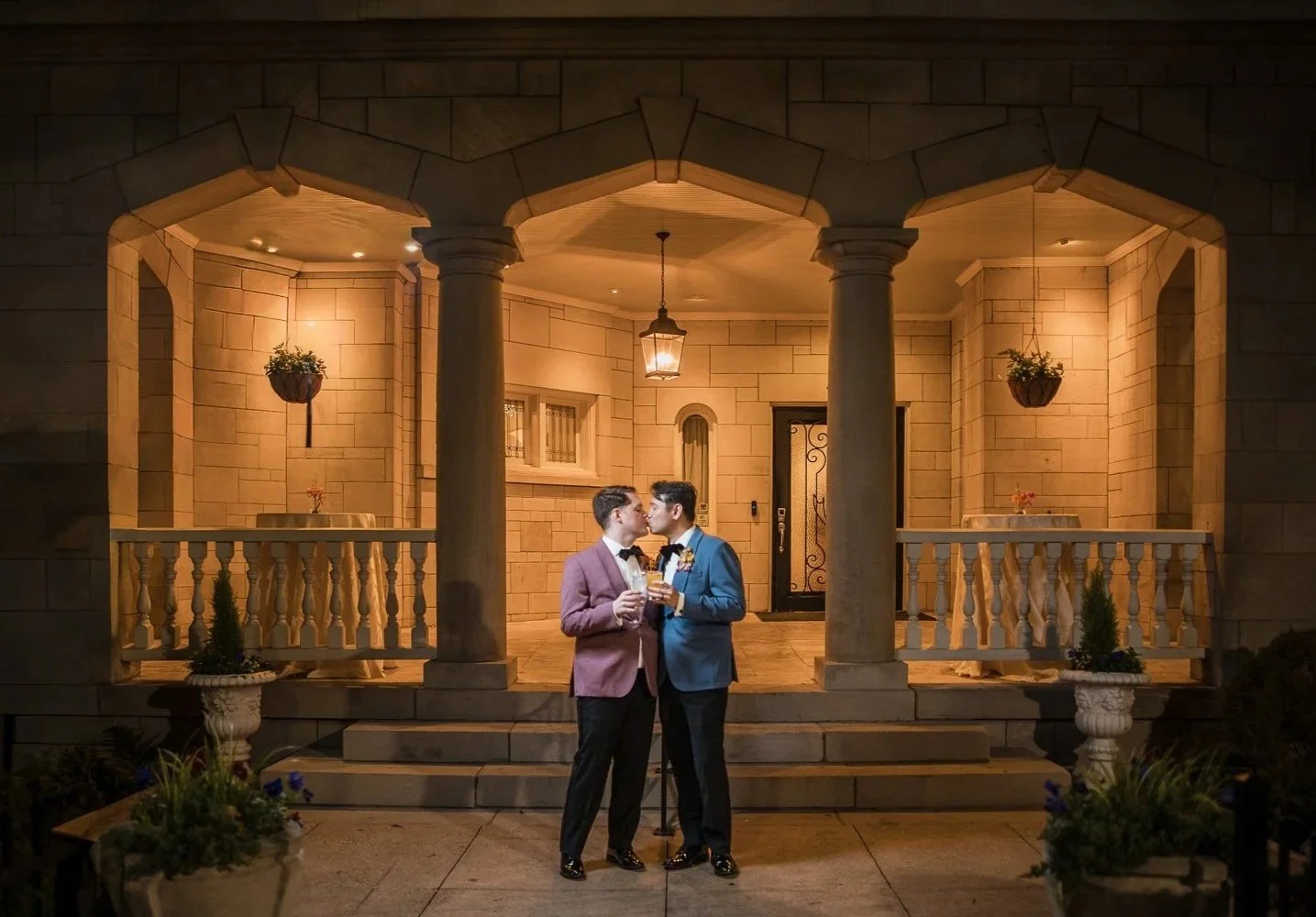 Two men in tuxedos kissing each other on the lips in front of a lit porch with stone columns, hanging flower baskets, and potted plants at night.