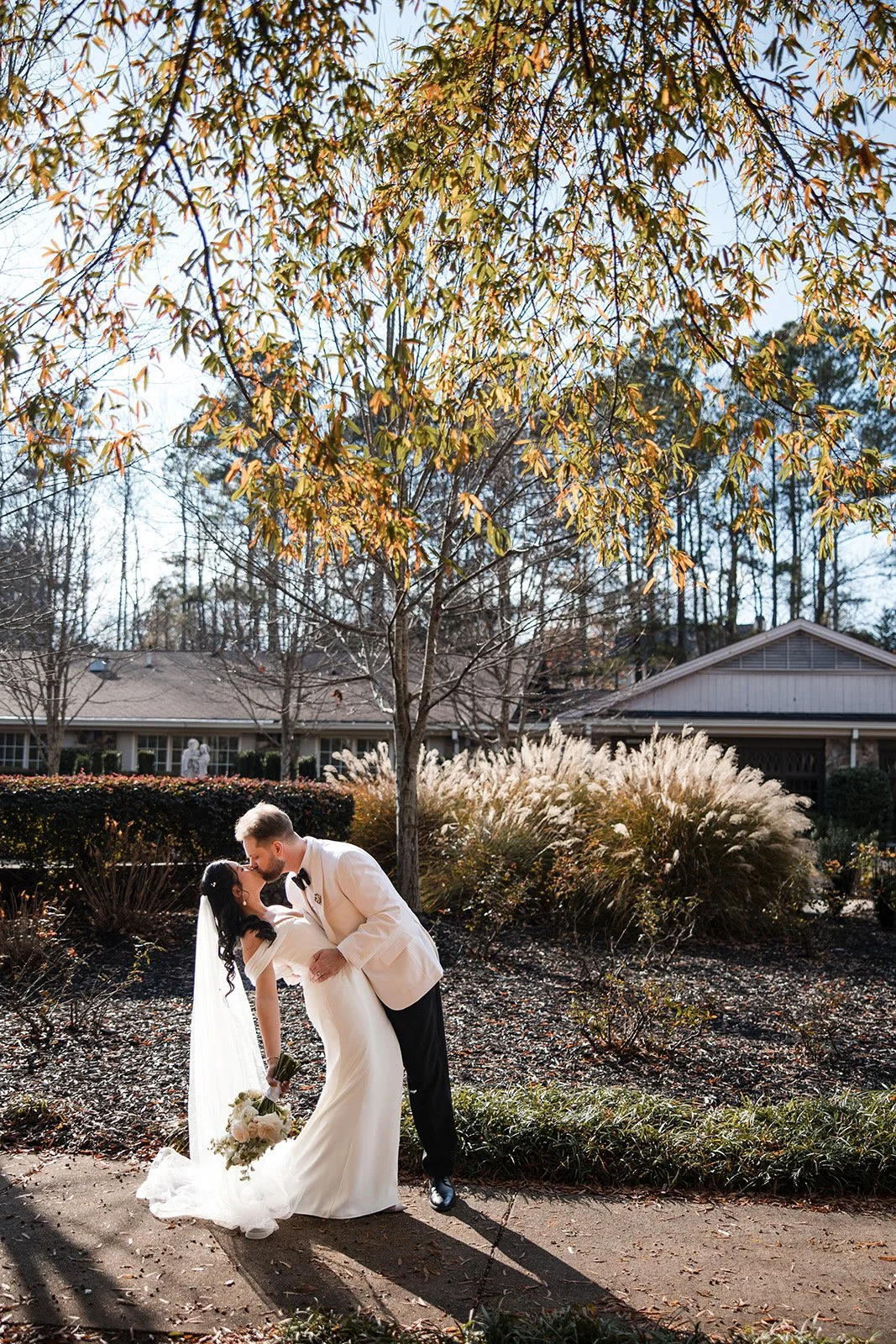 Couple in wedding attire sharing a romantic moment, embracing with eyes closed, outdoors near water with a railing in the background.