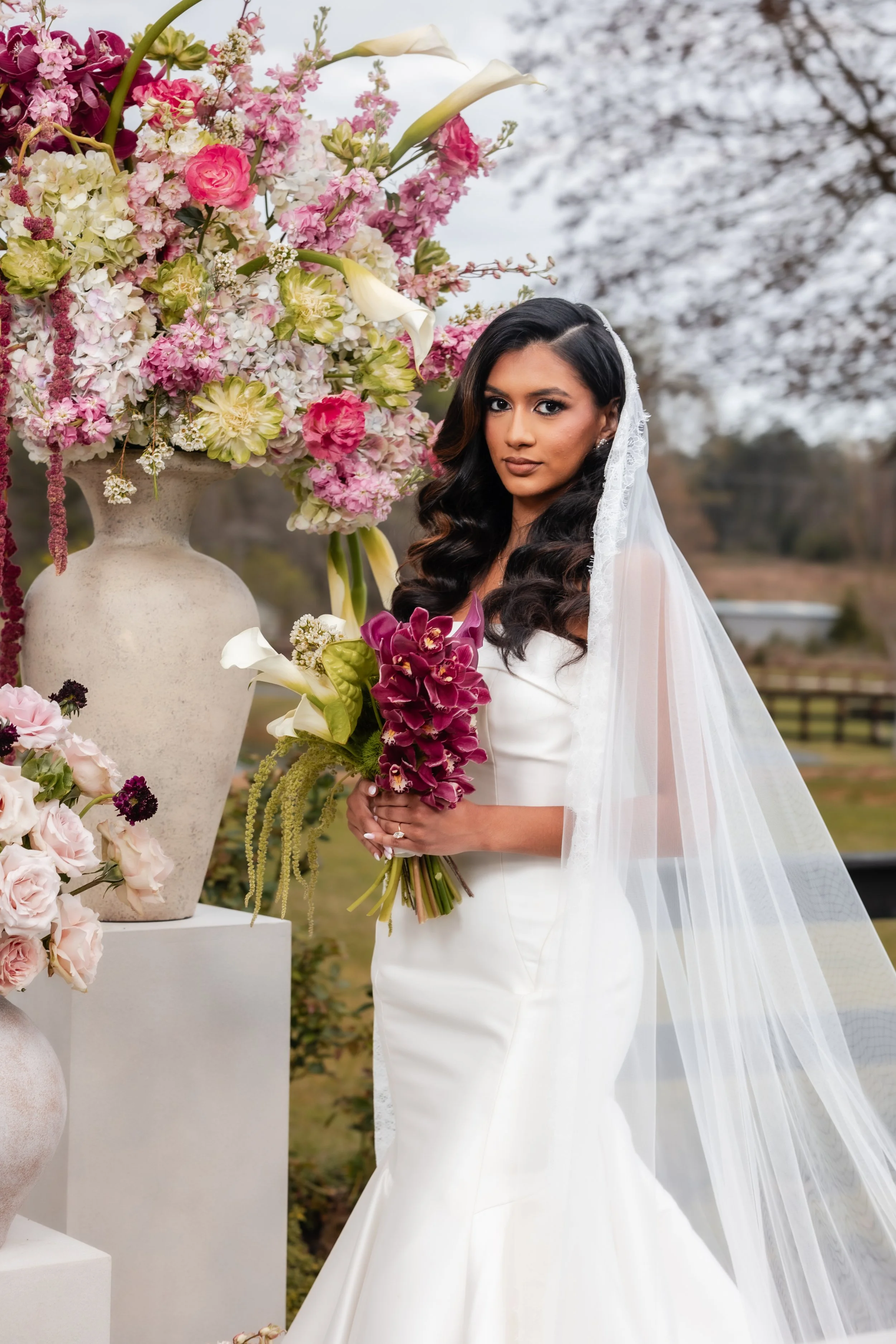 Bride in a strapless white wedding dress holding a large bouquet of pastel-colored flowers outdoors with trees and greenery
