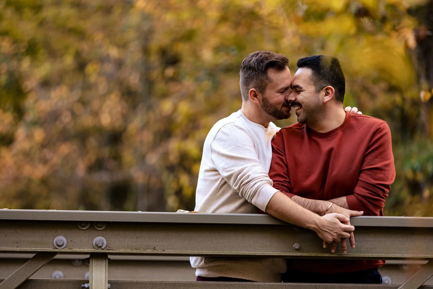 Two men are smiling and touching foreheads while leaning on a bridge railing in a park with autumn-colored trees in the background.