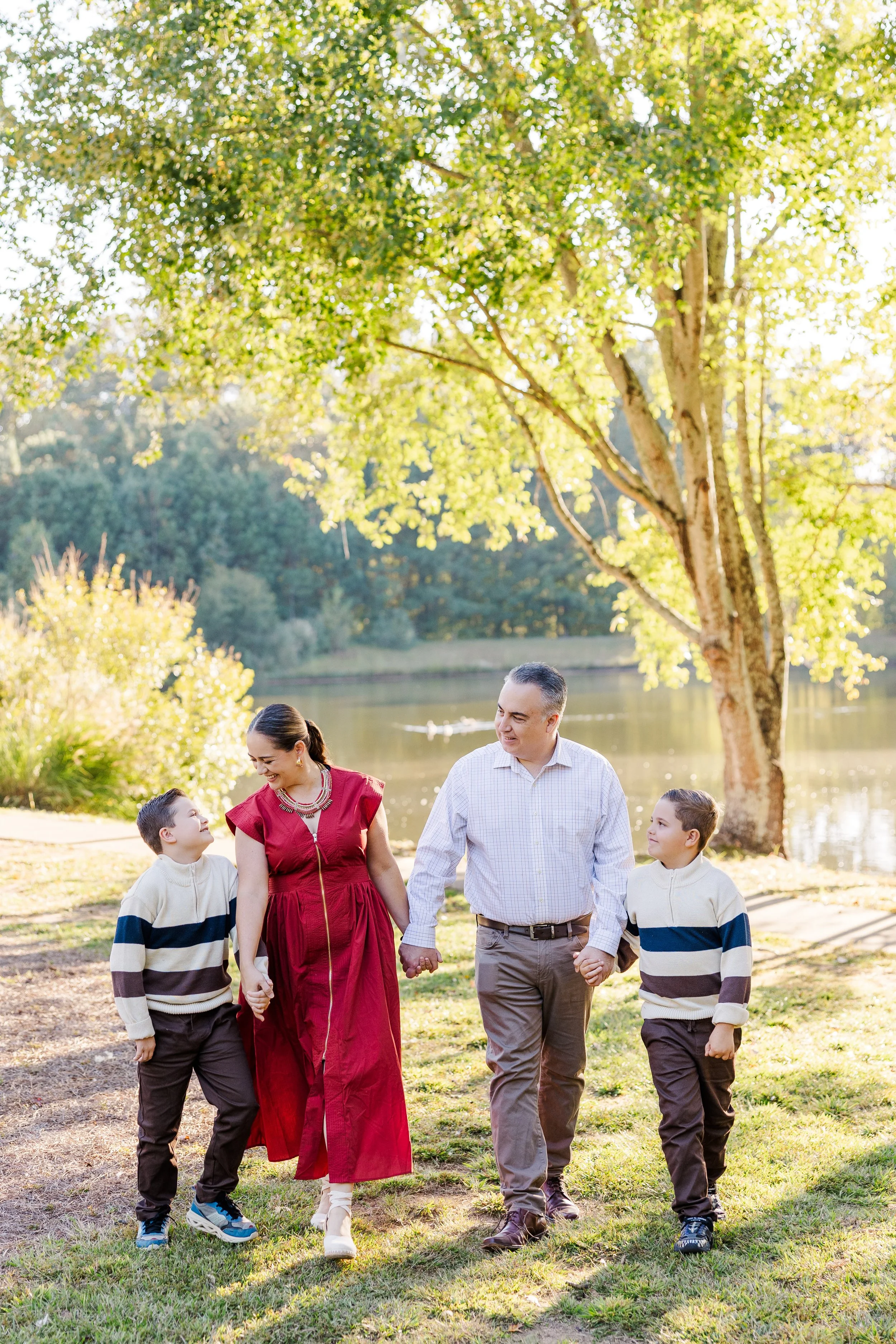 A family of four, two parents and two children, enjoying a walk by the lake surrounded by greenery and trees during the daytime.