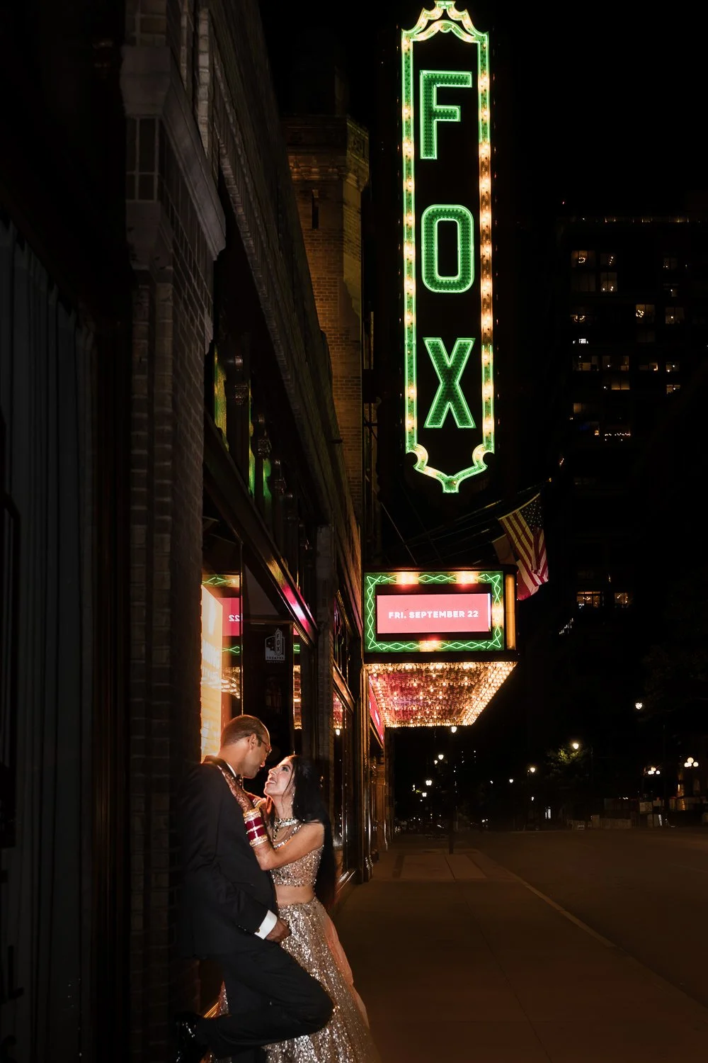 Indian Couple night photography outside the Fox Theater in Atlanta, dressed in Indian formal attire, illuminated by theater marquee lights, marquee sign displaying wedding date.