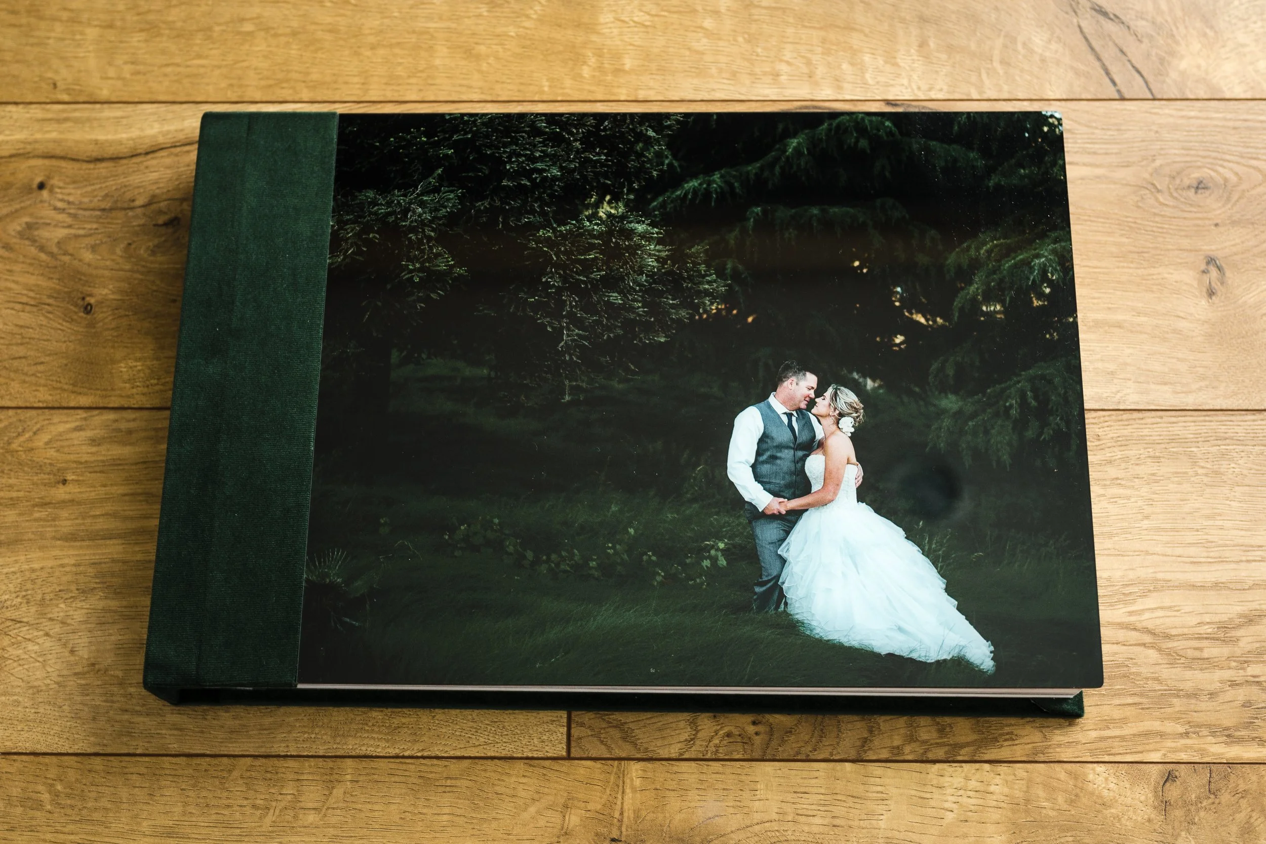 A hardcover photo album with a picture of a bride and groom on the cover, standing outdoors in front of trees, on a wooden surface.