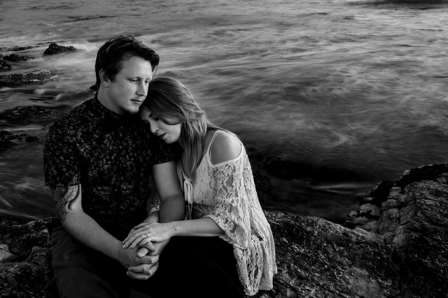 A black-and-white photo of a couple sitting on rocks by the ocean, with the woman resting her head on the man's shoulder, both holding hands, during sunset.