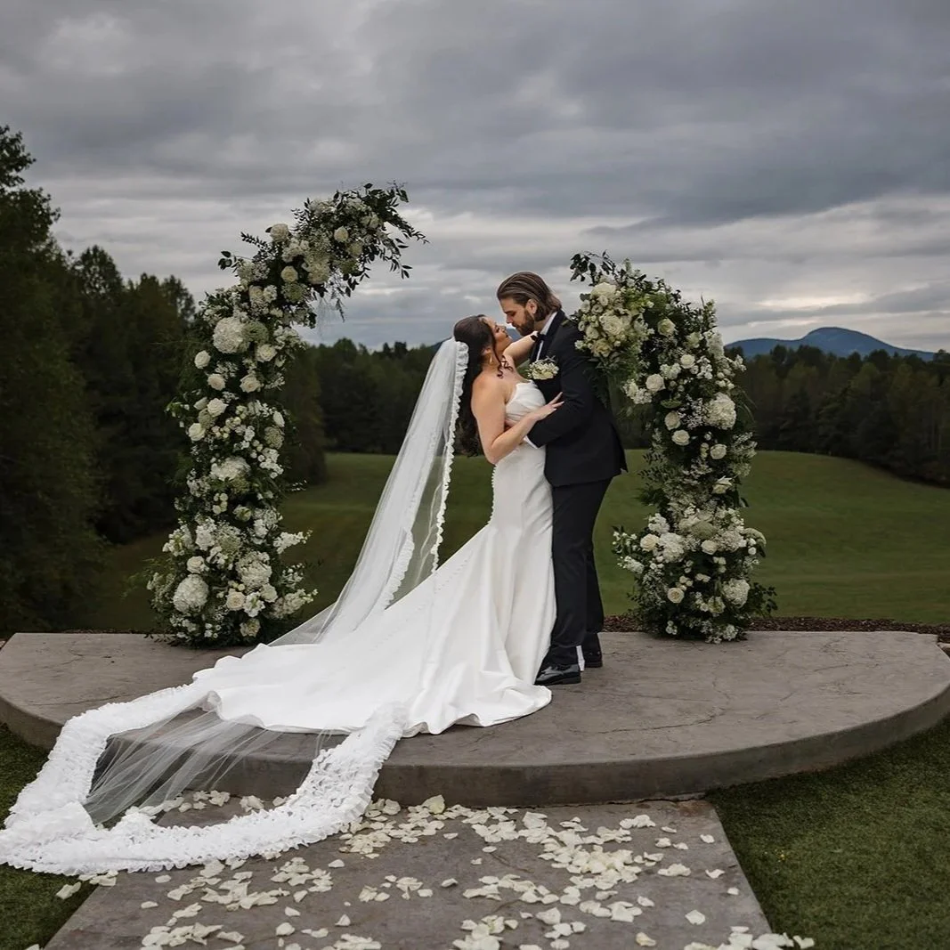 A bride and groom sharing a kiss while standing on a circular platform outdoors surrounded by a large floral arch of white flowers. The scene is set against a cloudy sky and scenic landscape.