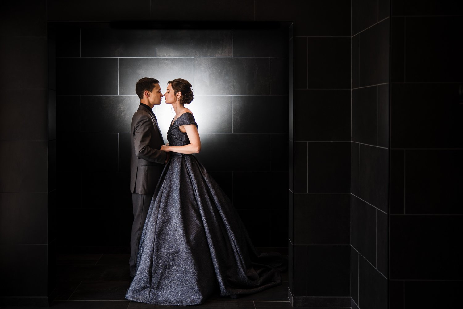 A man and woman in formal attire standing close, about to kiss, in front of a black tiled wall with backlighting.