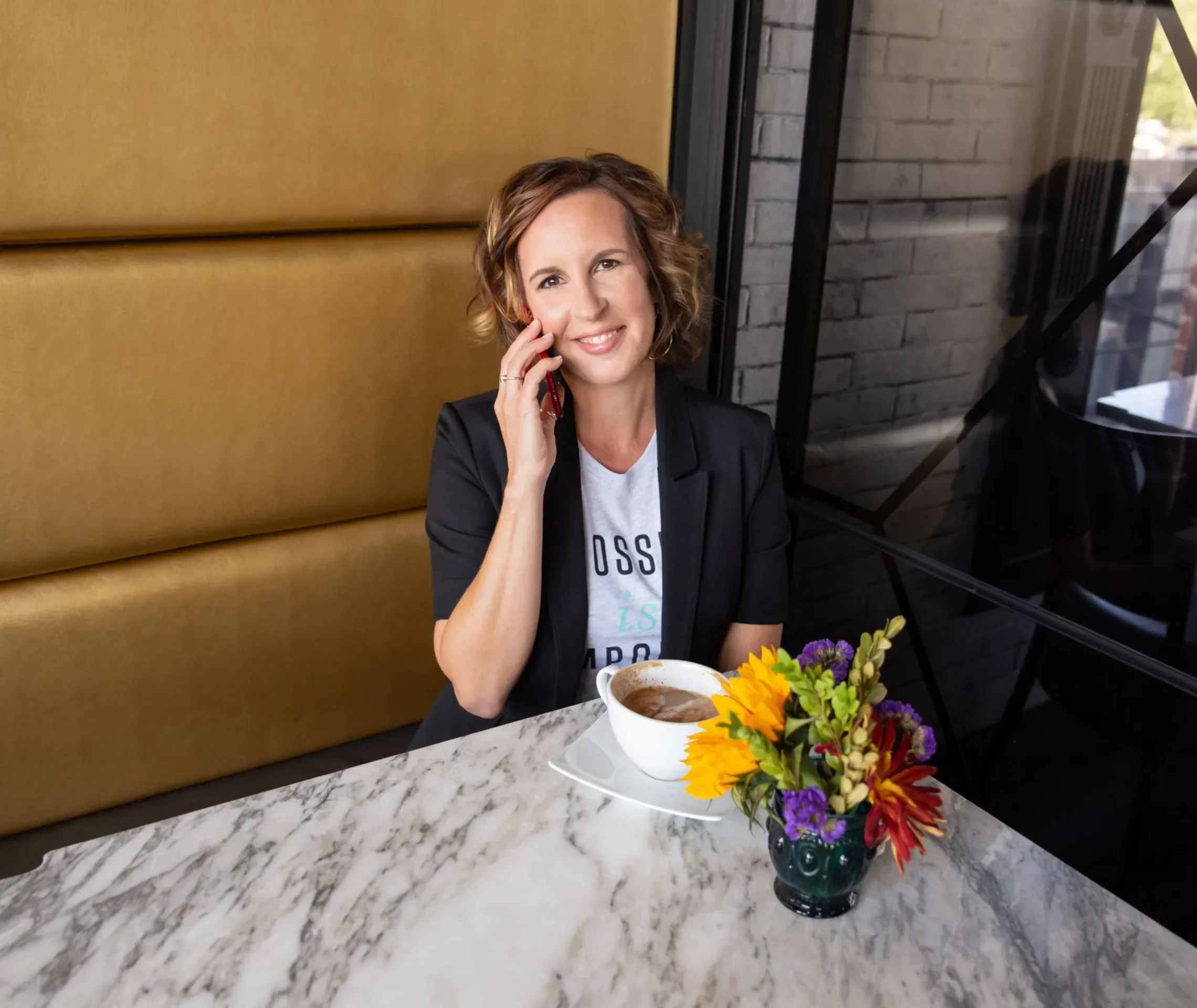 Voxer Coach Melanie Shmois with curly, shoulder-length brown hair, smiling while talking on a cell phone, seated at a marble table with a cup of coffee, a bouquet of colorful flowers, in a modern setting with yellow padded wall & exposed brick wall.