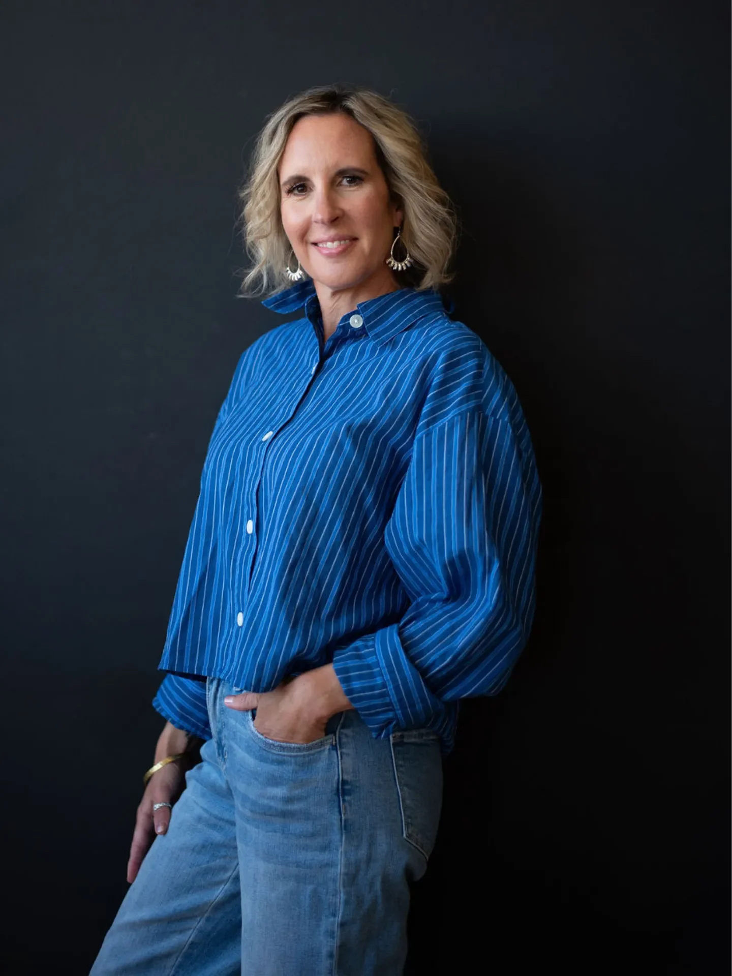 Learn about Executive High Performer Coach, Melanie Shmois, with blonde wavy hair wearing a blue striped button-up shirt and light blue jeans, standing against a black background, smiling softly.