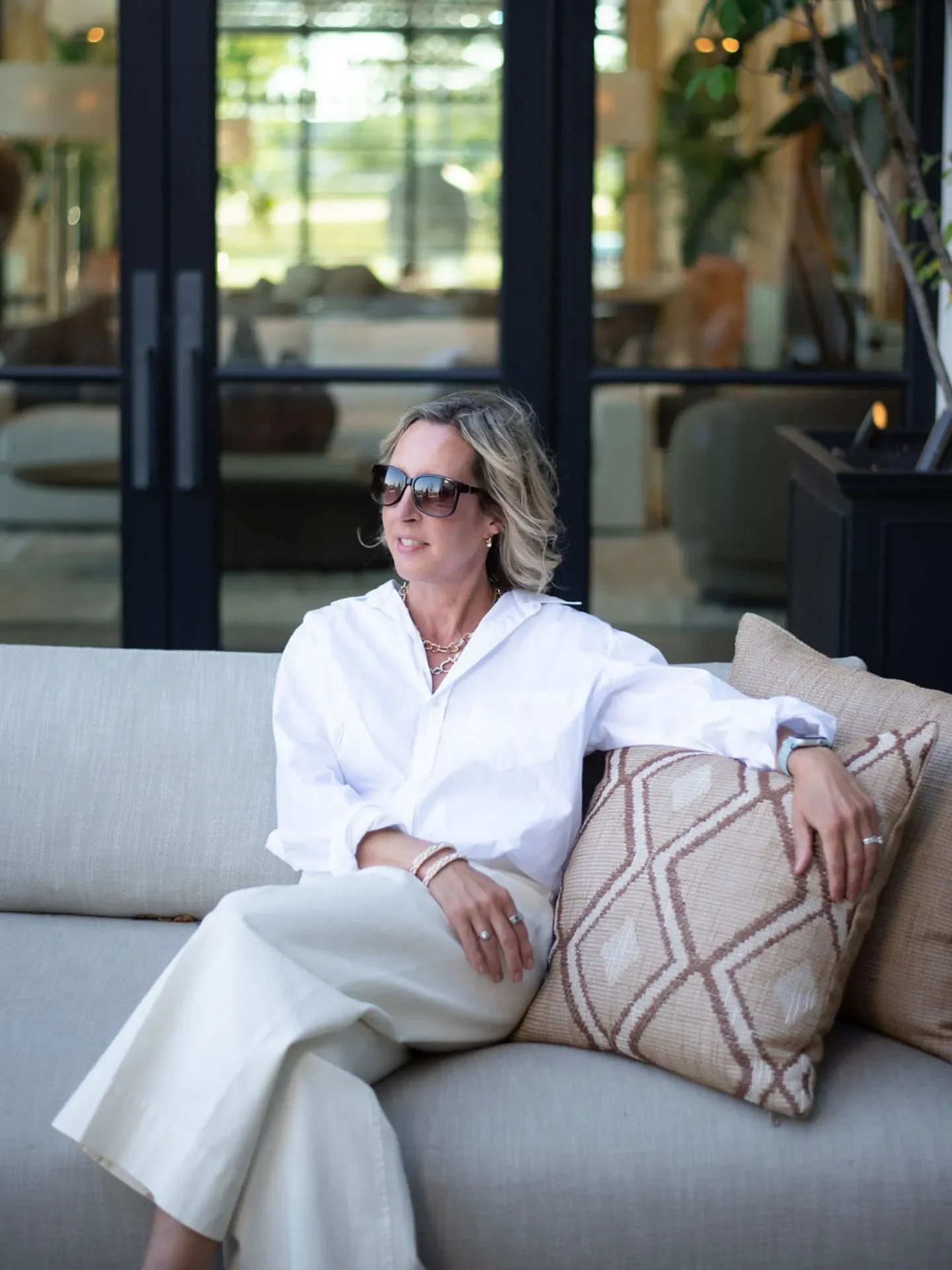 A woman with blonde hair, wearing sunglasses and a white shirt, is sitting on a light-colored sofa, leaning back with her arm resting on a patterned pillow.