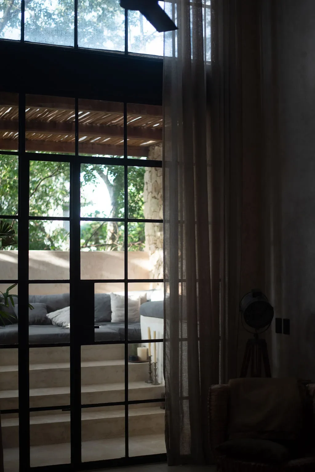 Indoor view through a glass door with black frame, revealing an outdoor patio area with steps, a cushioned seating area, and greenery with trees and plants.
