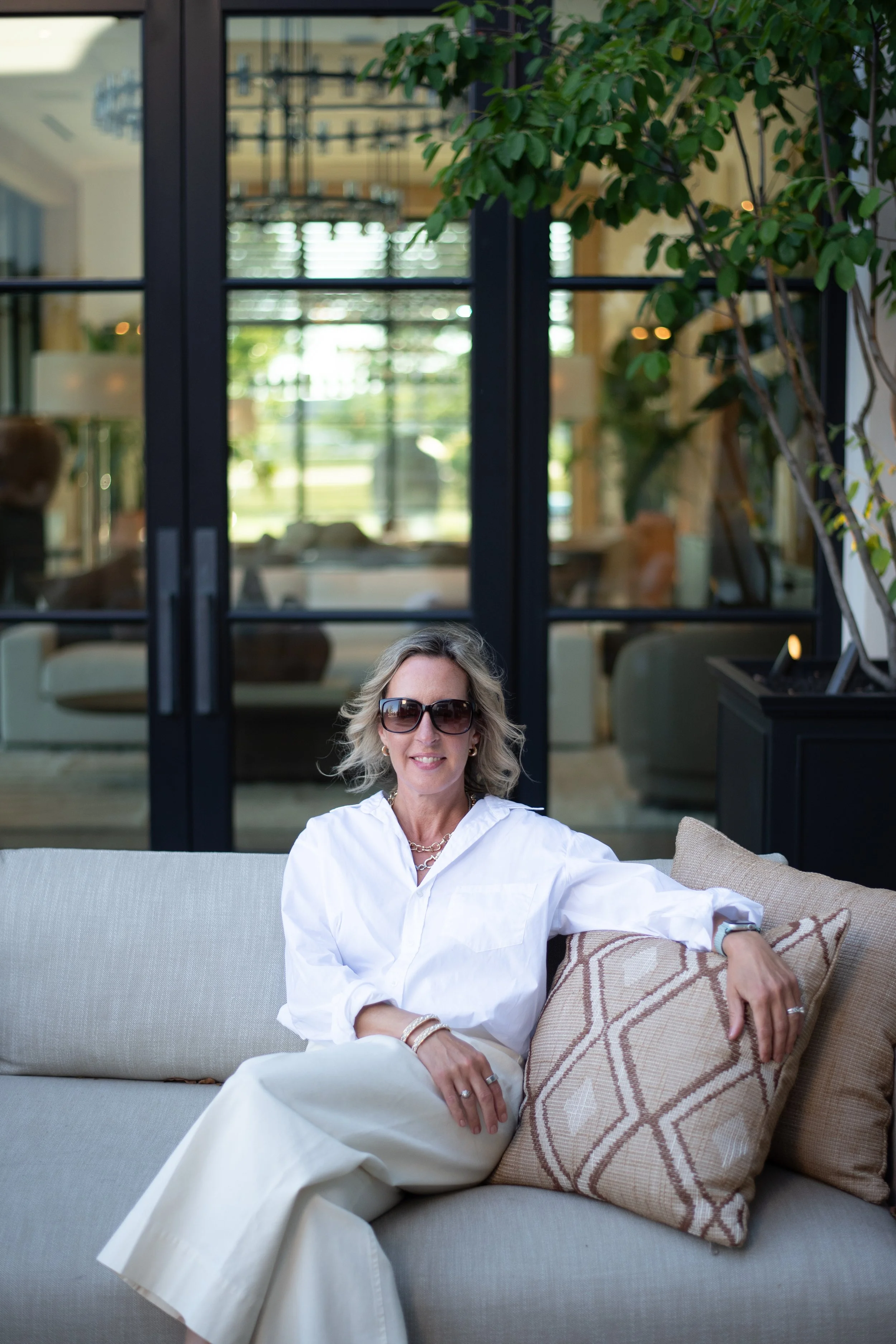 A woman with blonde hair wearing black sunglasses, a white blouse, and cream-colored pants is sitting on a beige outdoor sofa, relaxed, with her left arm resting on a patterned pillow. She is smiling and looking at the camera. The background shows large black-framed glass doors and a lush green indoor plant.
