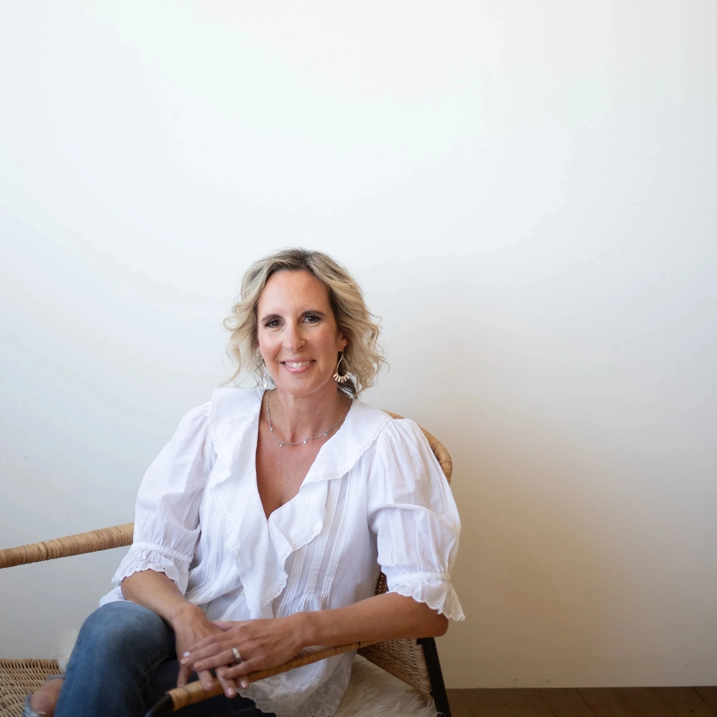 A woman with blonde, curly hair and earrings sitting on a wicker chair, smiling, wearing a white blouse, in front of a plain white wall.
