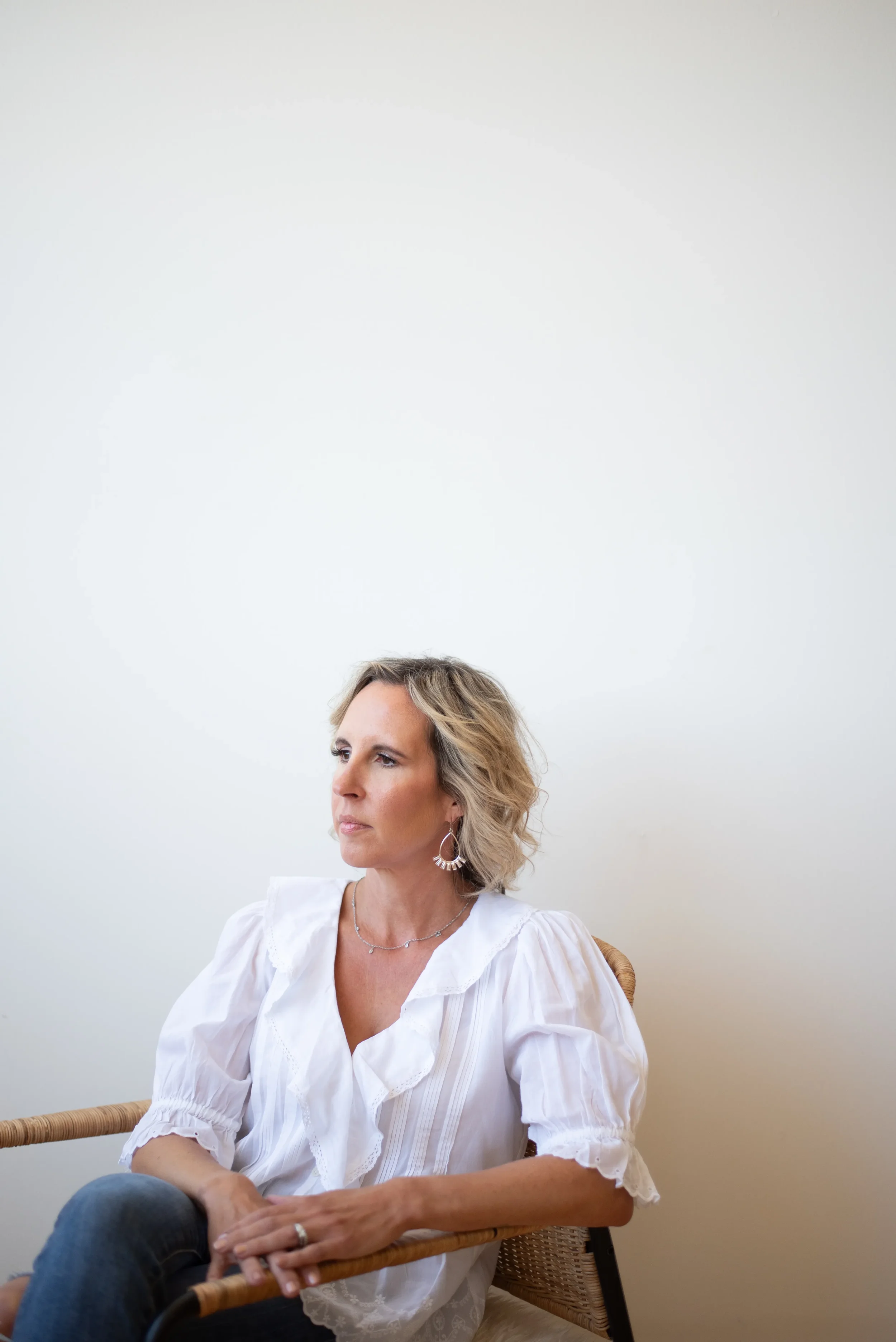A woman with blonde, shoulder-length hair, wearing a white blouse, sitting in a wicker chair with a light-colored wall behind her. She is looking to her left with a thoughtful expression, holding her left arm with her right hand.