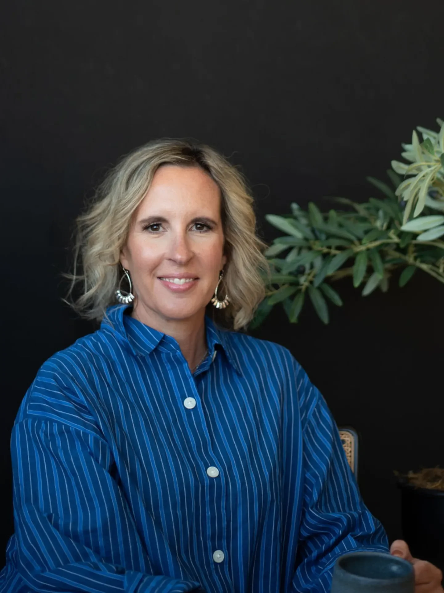 A woman with blonde hair in a blue pinstripe shirt sitting in front of a black background with a green plant on the right side, holding a mug.