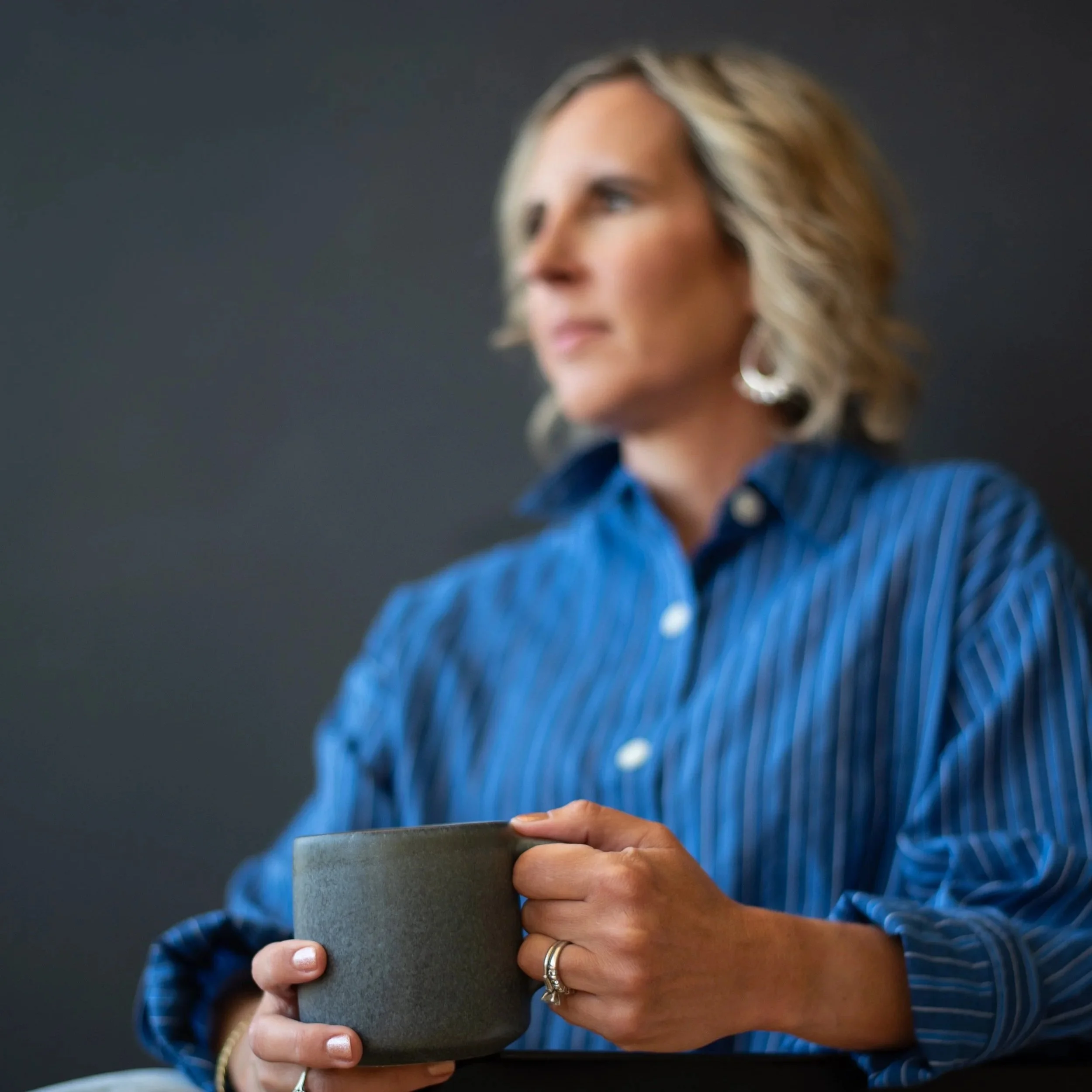 Close-up of a woman's hand holding a gray coffee mug, with a blurred woman in the background wearing a blue shirt, and dark solid-colored background.