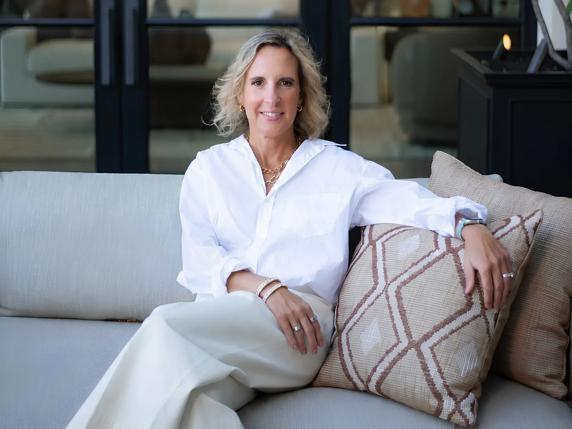 Melanie Shmois - Personal Coach for High Performers sitting outdoors on a couch with brown accent pillows wearing a white blouse and off-white pants with her arm on her crossed legs
