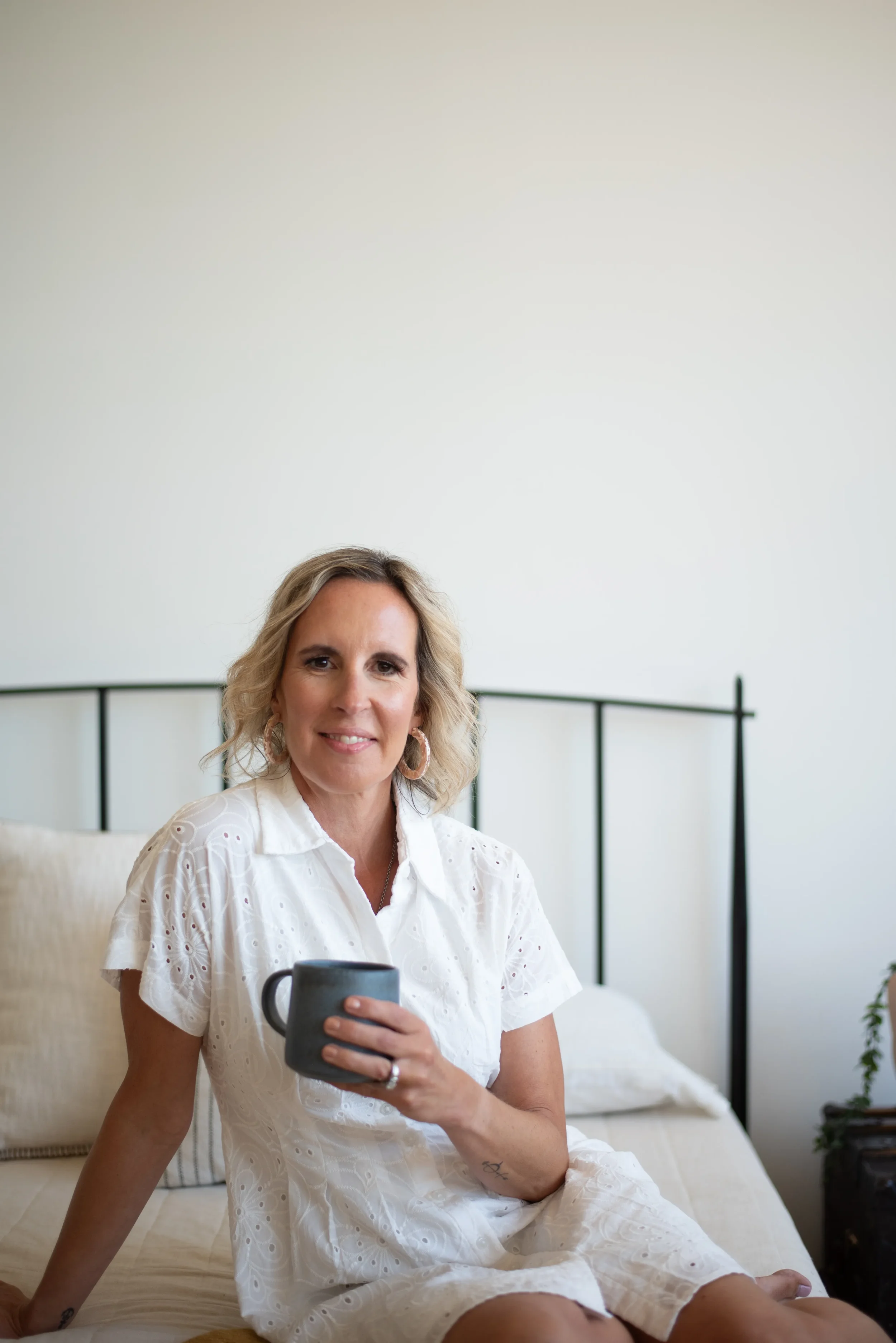A middle-aged woman with blonde hair and hoop earrings is sitting on a bed, holding a gray mug, and smiling at the camera in a bright, minimal bedroom with white walls and a black metal bedframe.