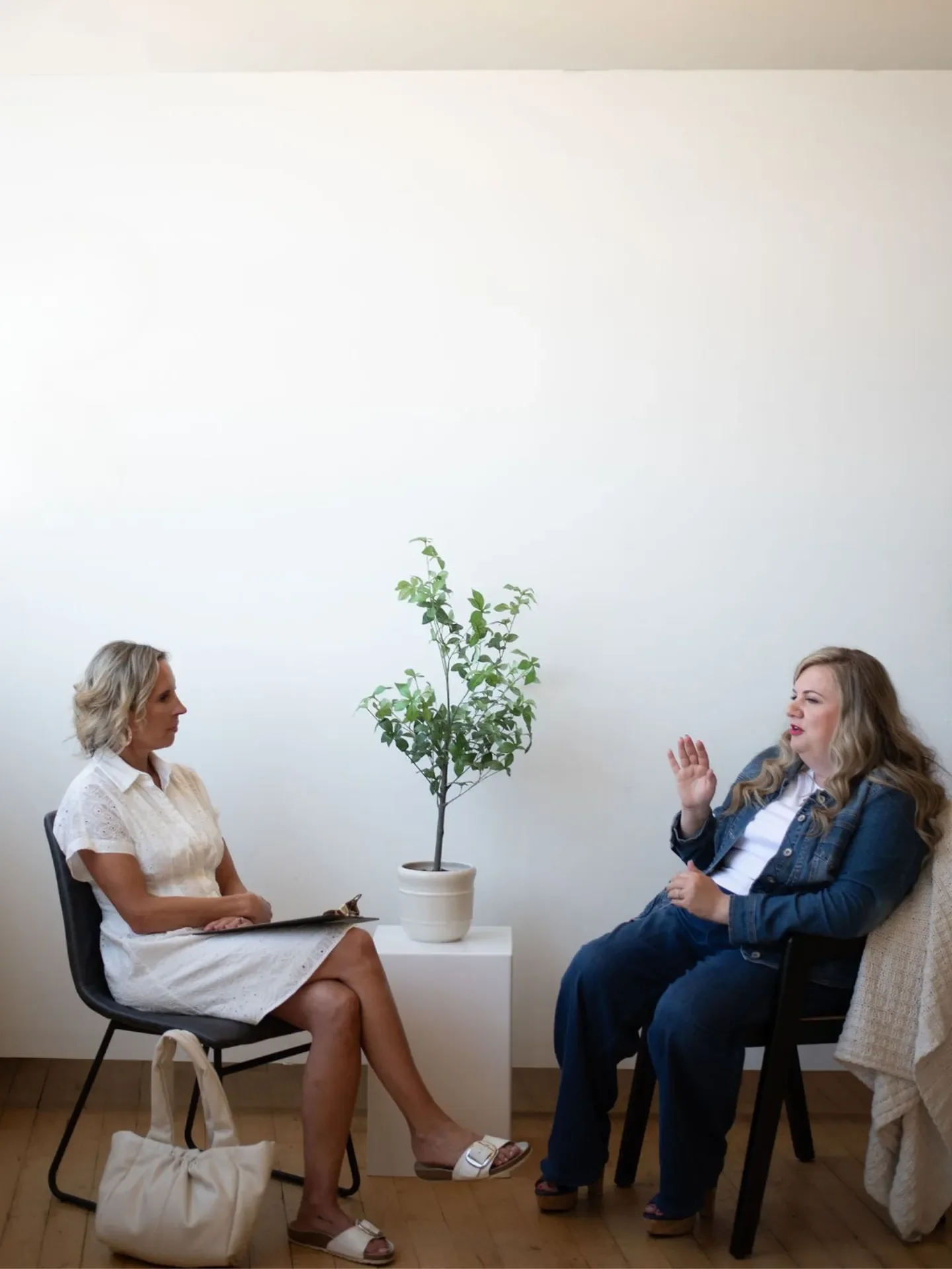 Two women are sitting across from each other having a conversation in a minimalistic room with white walls. There is a potted plant in between them on a small white table, and a tote bag on the floor beside one woman. The woman on the left has short blonde hair, is wearing a white dress and sandals, and is holding a tablet. The woman on the right has long wavy hair, is dressed in a denim jacket, white shirt, and jeans, and is gesturing with her hand.