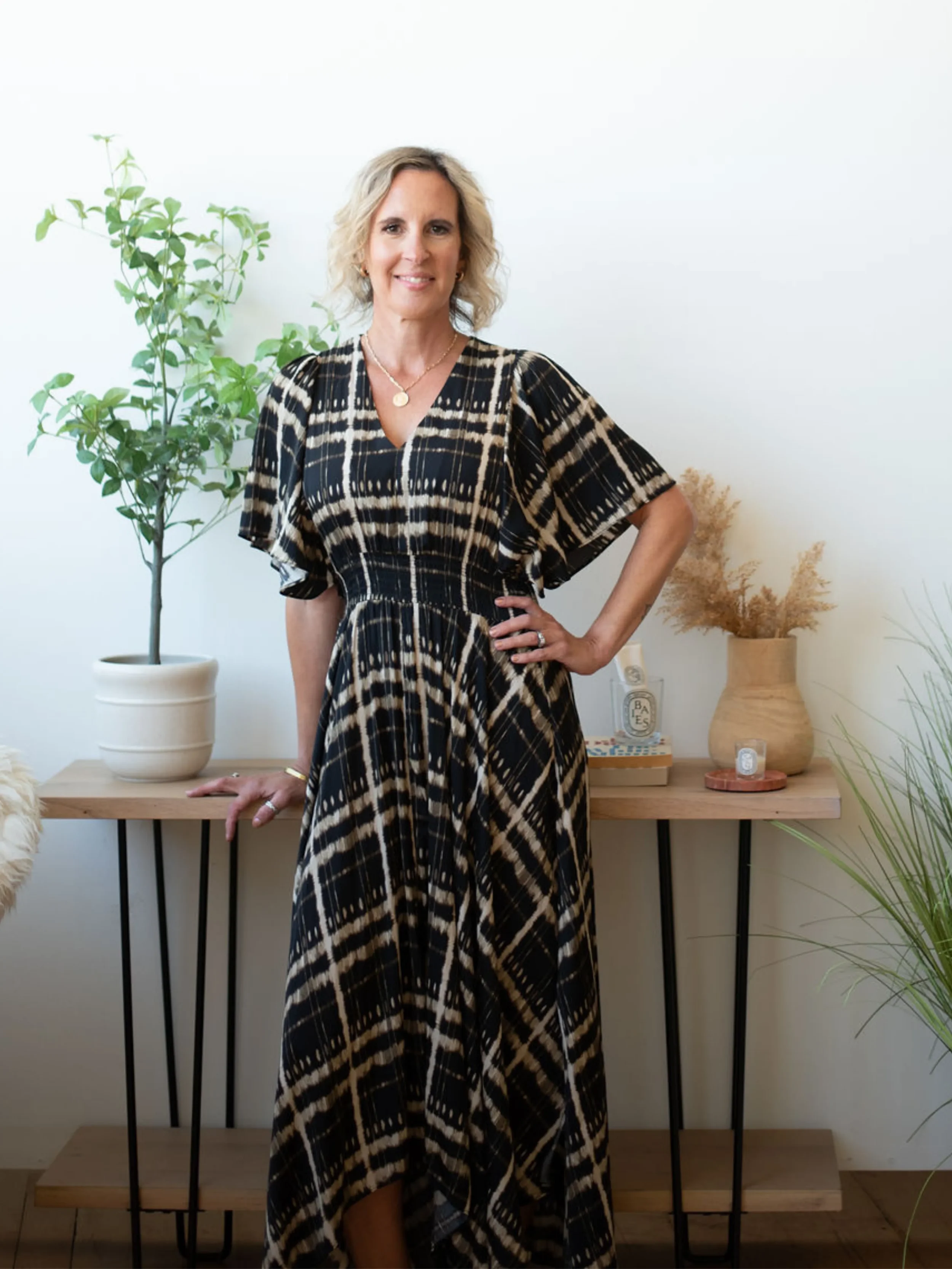 A woman with blonde hair wearing a black and beige patterned dress, standing in front of a table with decorative items and greenery in a bright room.