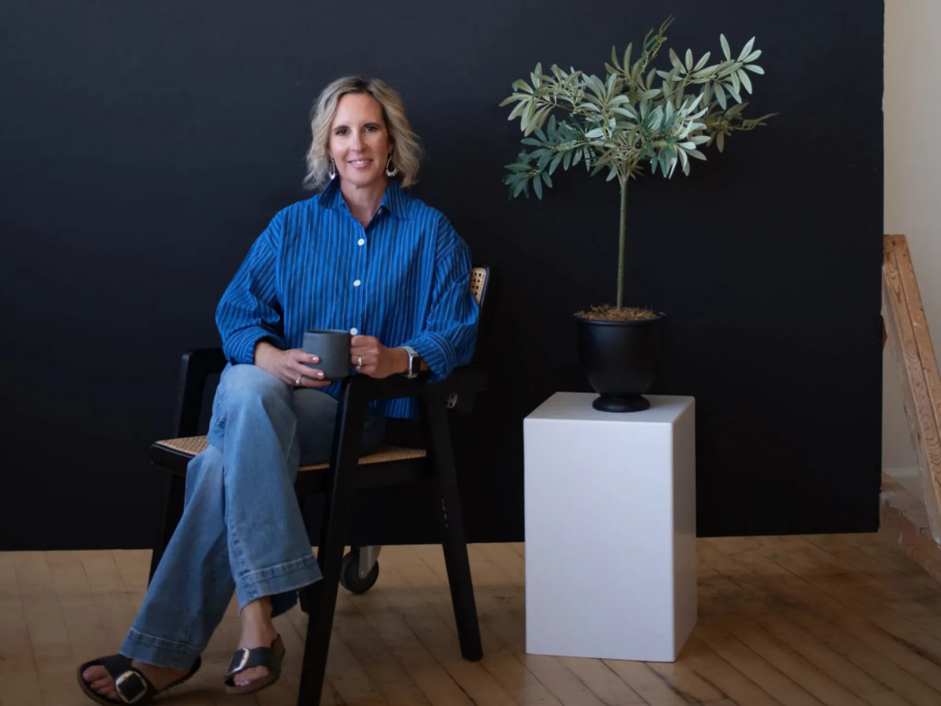 A woman with blonde, curly hair, wearing a blue striped shirt and jeans, sits on a chair holding a cup, with a potted tree and a black wall in the background.