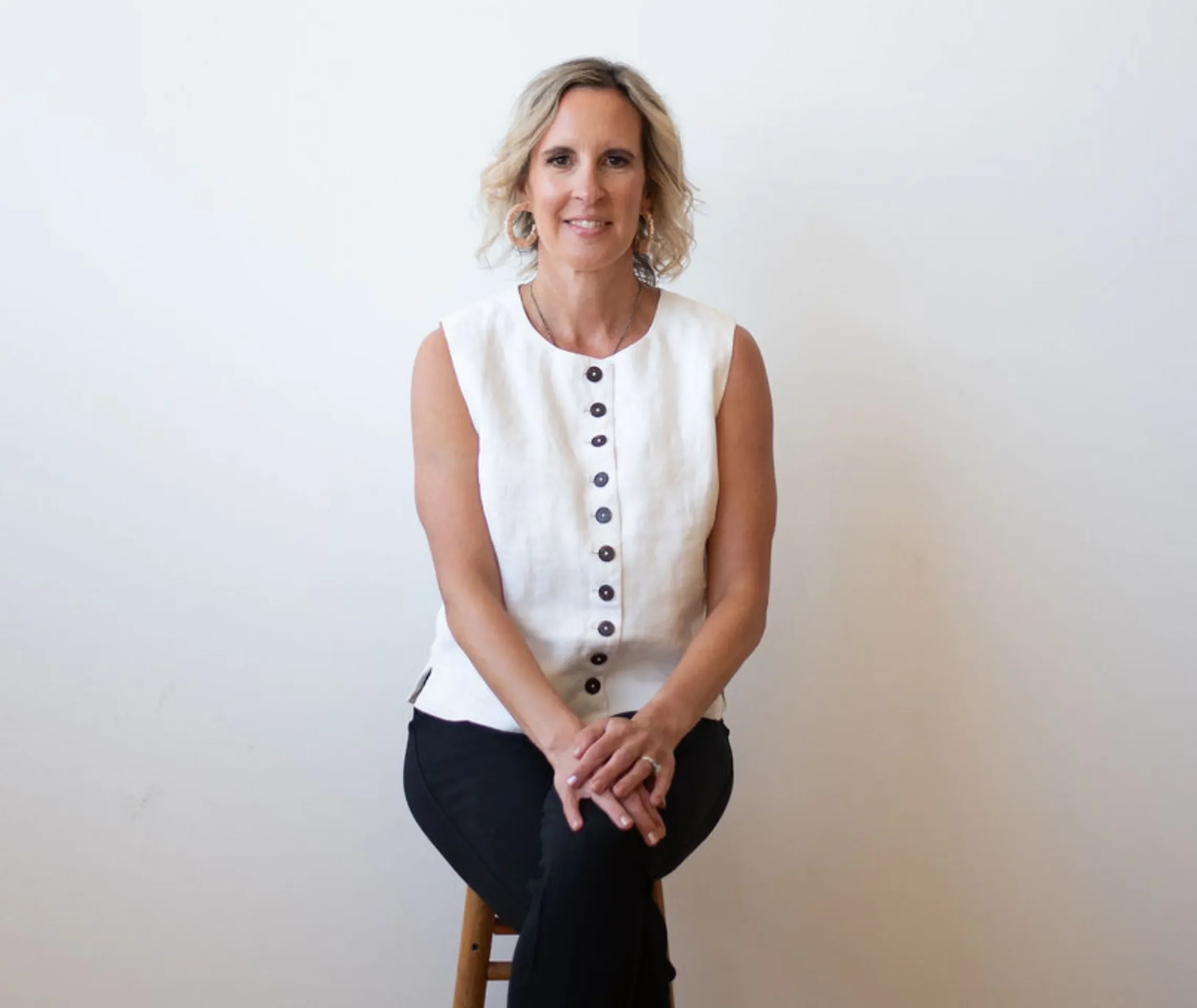 A woman with wavy blonde hair smiling, wearing large hoop earrings, a sleeveless white button-down top, and a black skirt, sitting on a wooden stool against a plain white wall.
