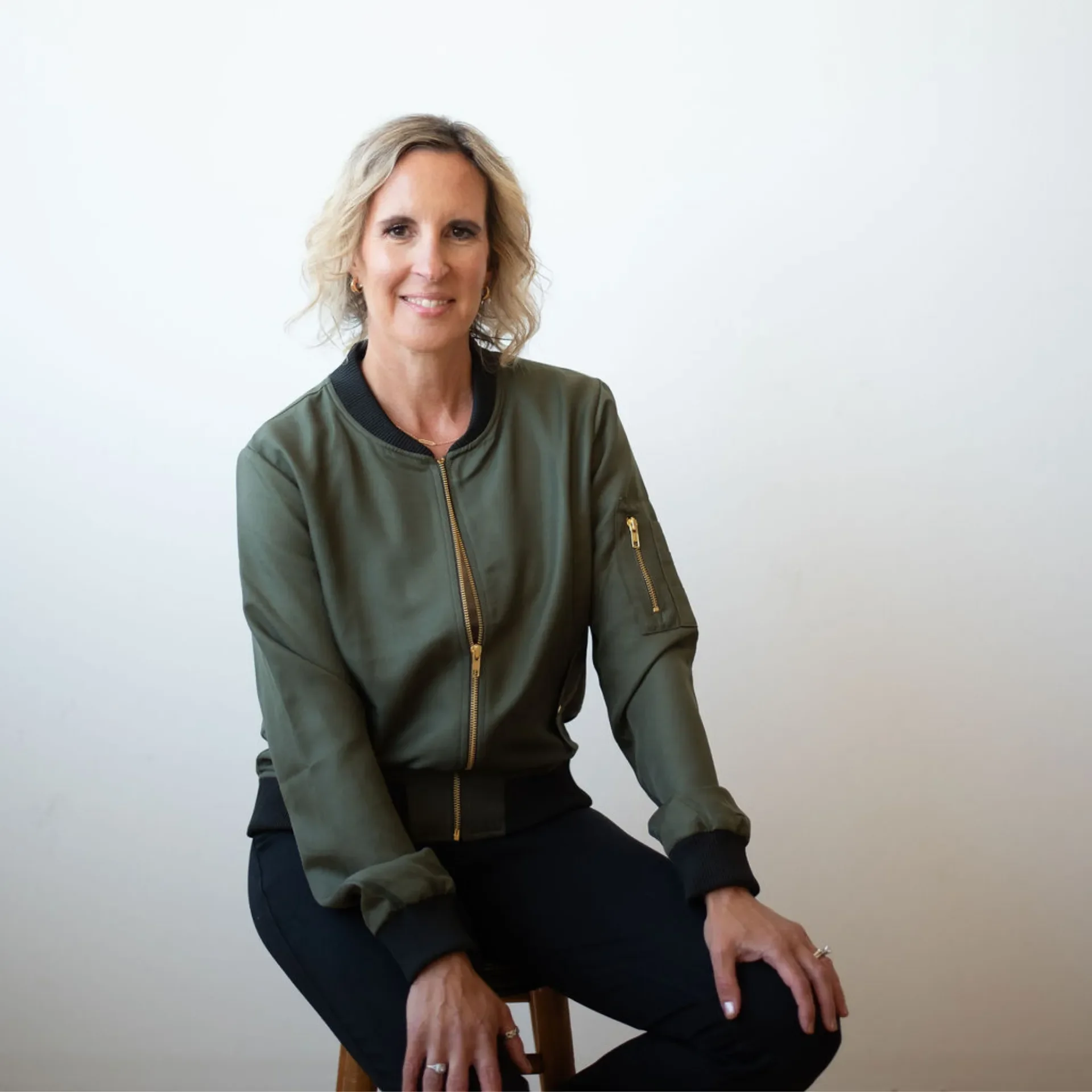 Media request for Personal Coach Melanie Shmois is wearing a green bomber jacket and black pants, sitting on a wooden stool against a plain white wall.