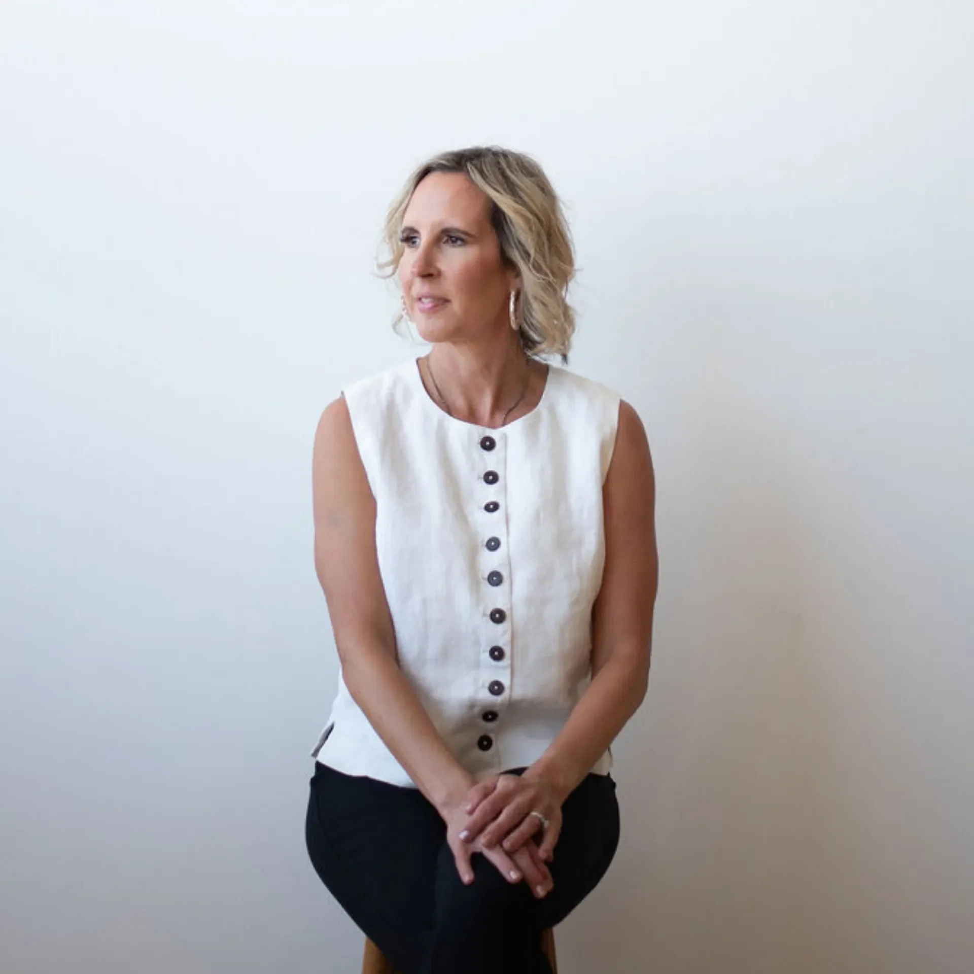 A woman with short blonde hair wearing a sleeveless white button-up shirt and black pants standing against a plain white wall.