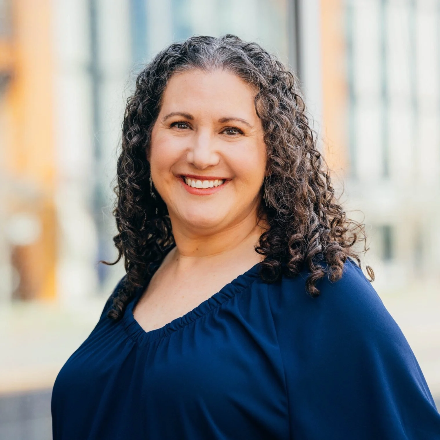 Portrait of a smiling woman with curly hair, wearing a navy blue top, standing outdoors near a building.