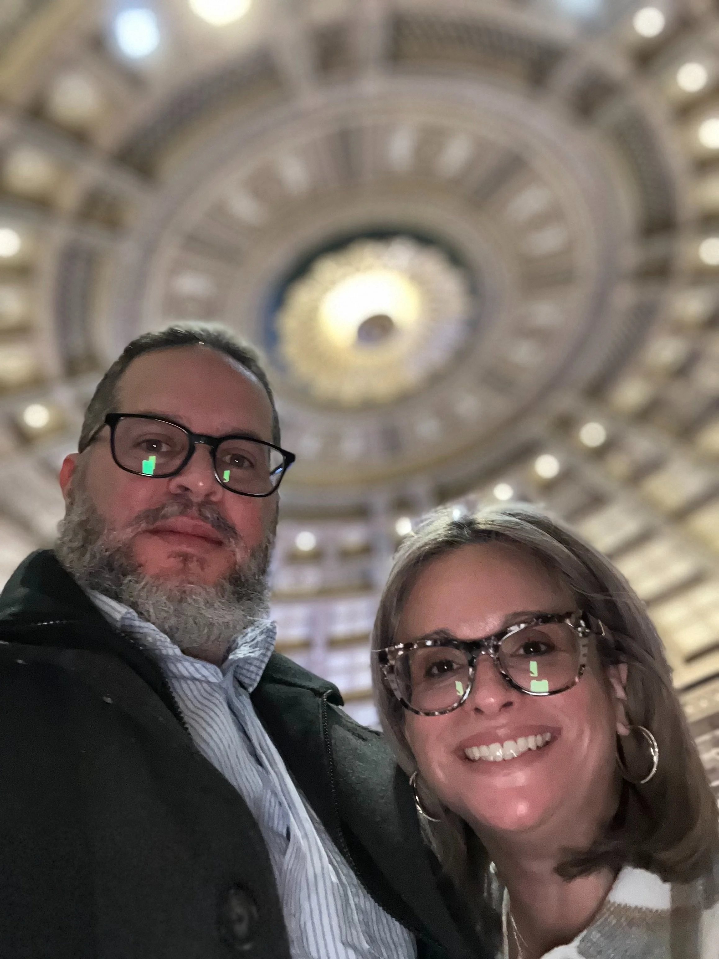 A man and woman taking a selfie inside a grand, circular building with a decorative, illuminated ceiling.