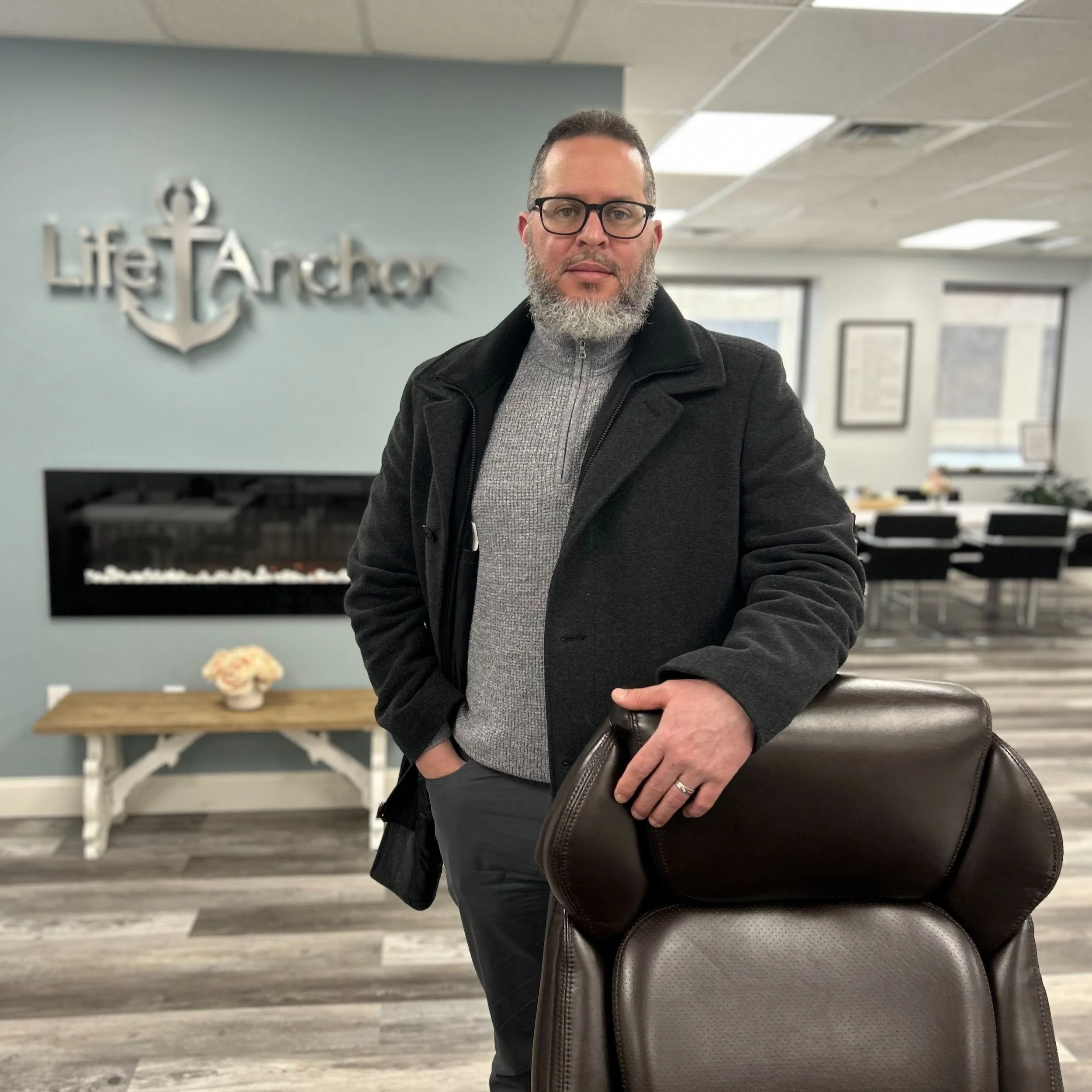 A man with glasses and a beard standing in an office, holding a brown leather chair. Behind him is a wall with a silver anchor and the words 'Lif t Anchor' and a fireplace. There is a small table with flowers on it and large windows with a view outside.