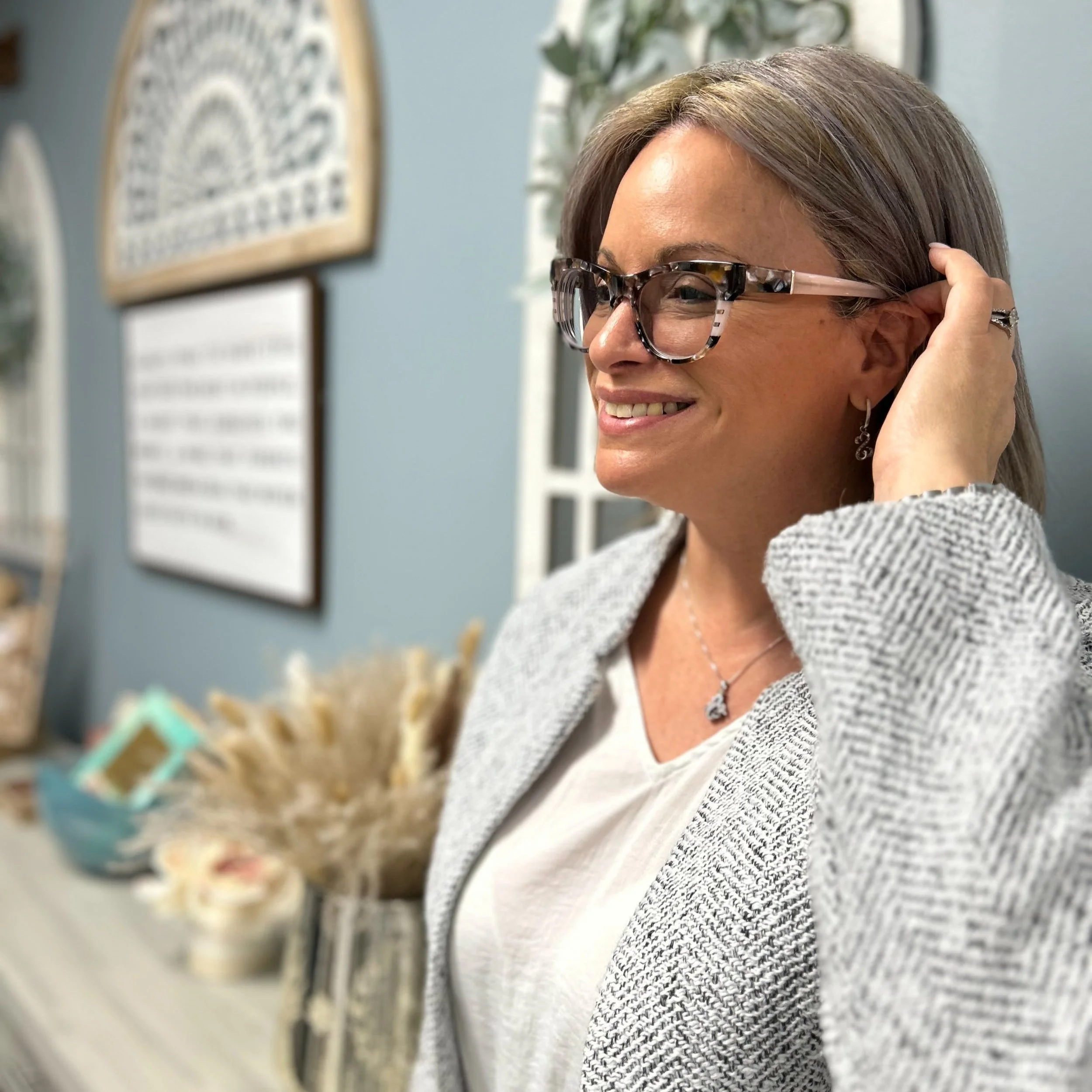 A woman with shoulder-length brown hair, wearing glasses, earrings, a necklace, and a gray blazer, smiling and touching her hair in a decorated indoor setting.