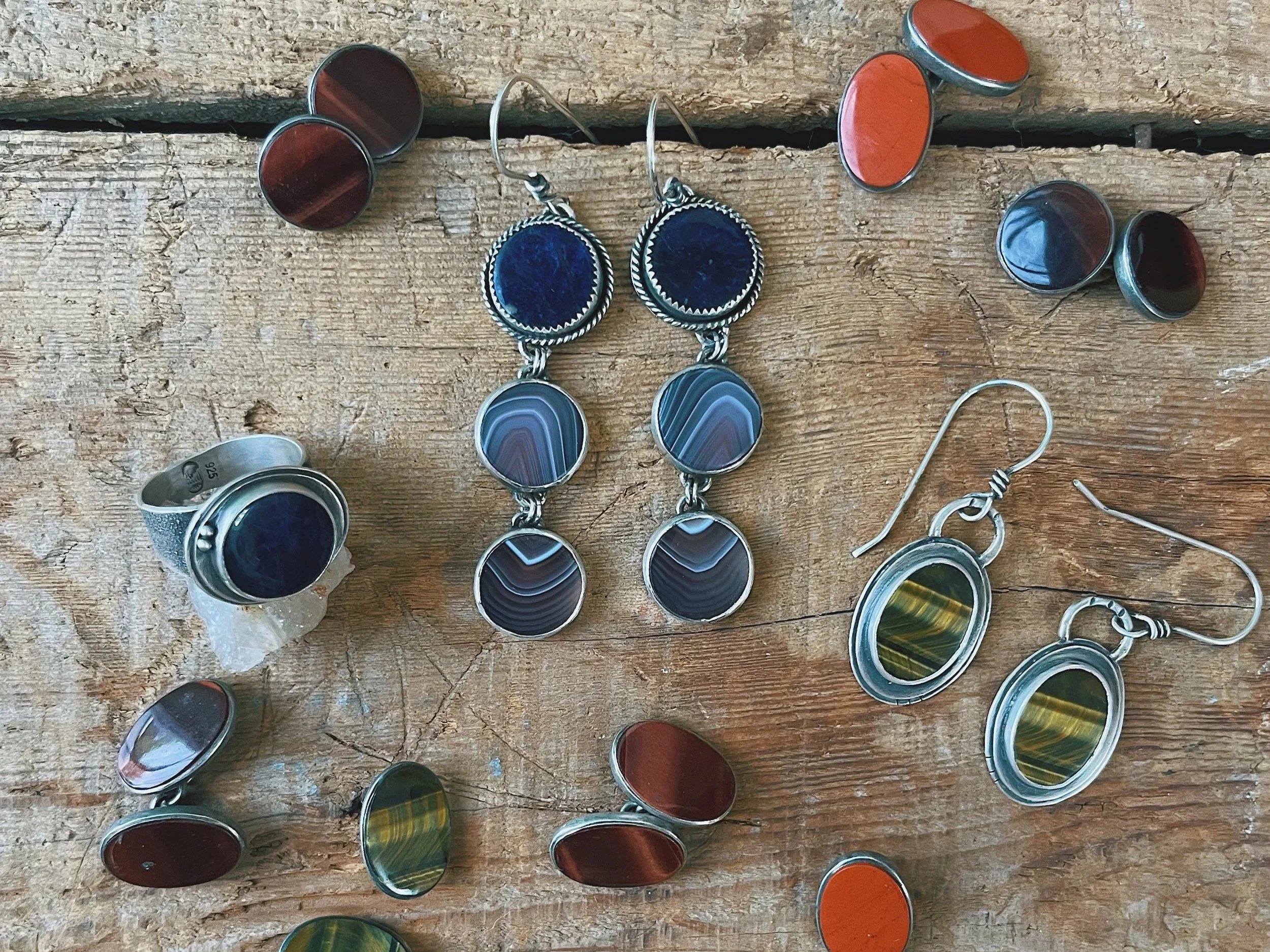 Various jewelry pieces including earrings, rings, and loose stones with a mix of colorful and metallic finishes, arranged on a wooden surface.