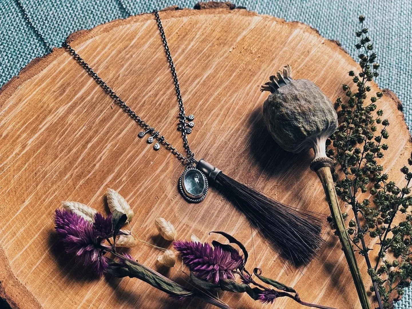 A decorative necklace with a gemstone pendant and black tassel, lying on a wooden surface with dried purple and beige flowers, a dried seed pod, and a sprig of greenery.
